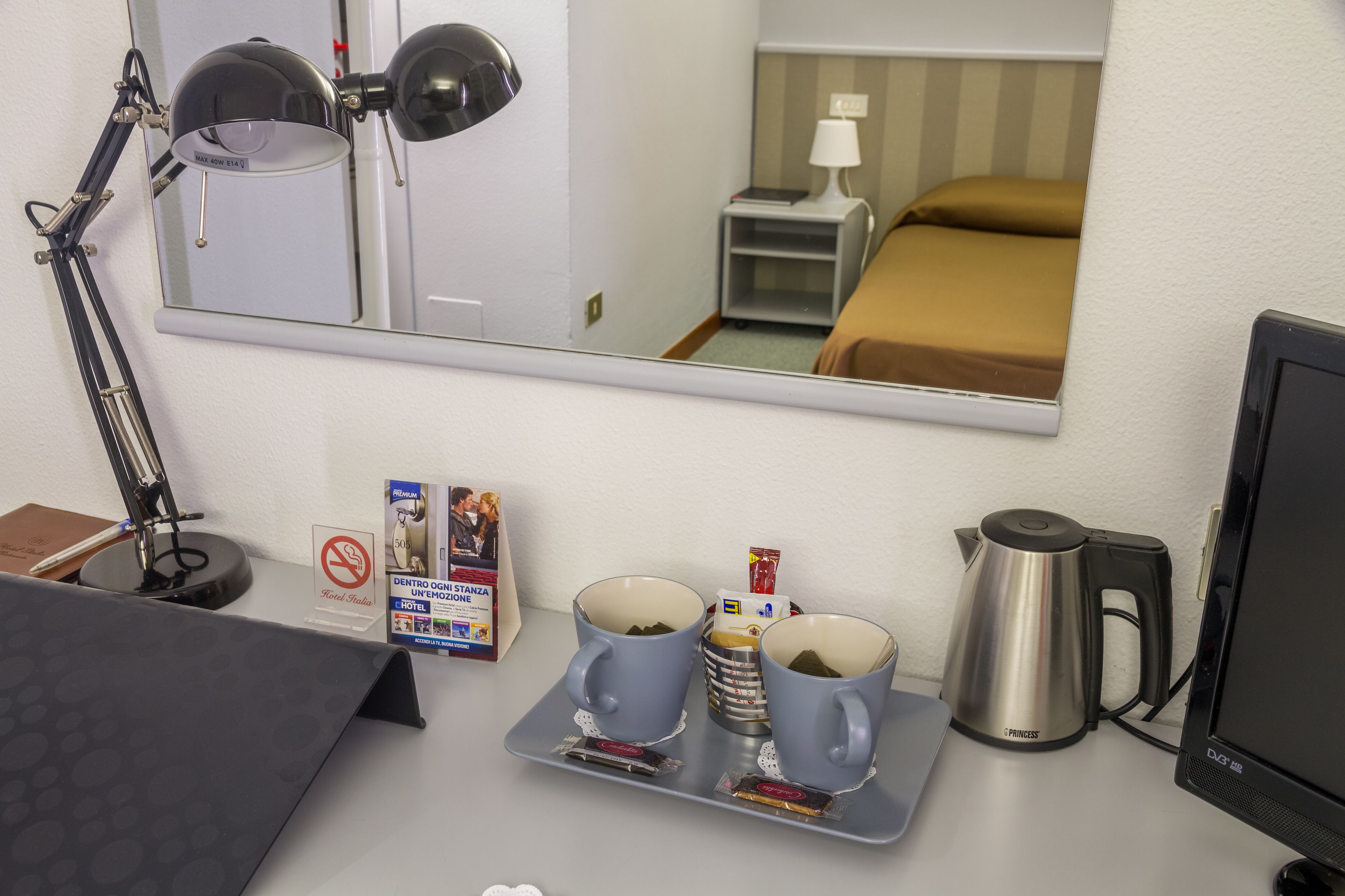 A hotel room with a desk, a mirror, a bed with a brown bedspread, a nightstand with a lamp, a coffee maker, and a tray with two mugs and a box of tissues.
