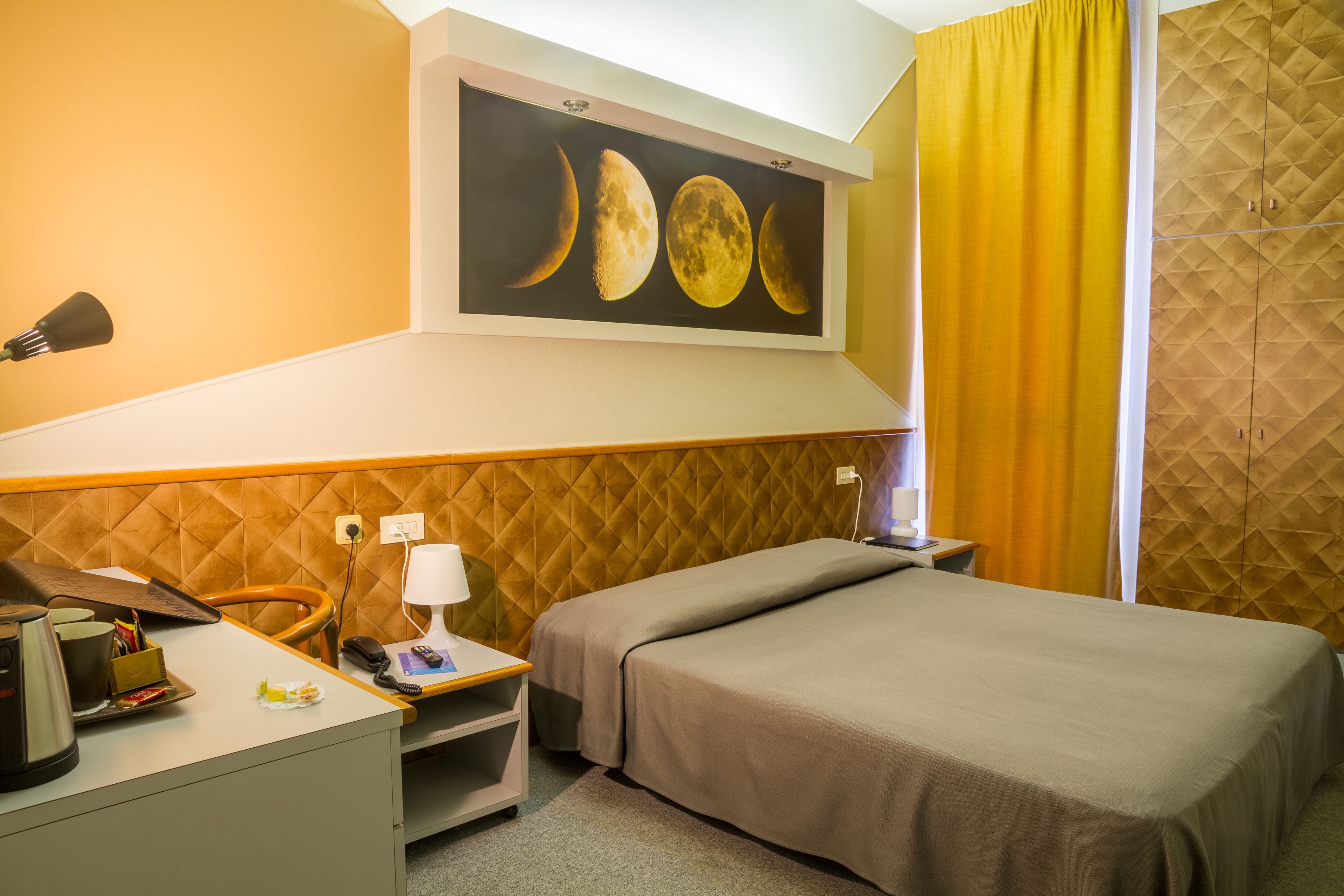 Una camera d'albergo con un letto, una scrivania con una lampada, una sedia, un quadro raffigurante le fasi lunari e una tenda gialla.