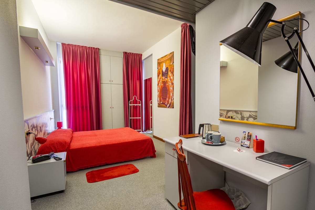 Habitación de hotel con dos camas, un escritorio, una silla, una lámpara de escritorio, un espejo y una cortina roja.