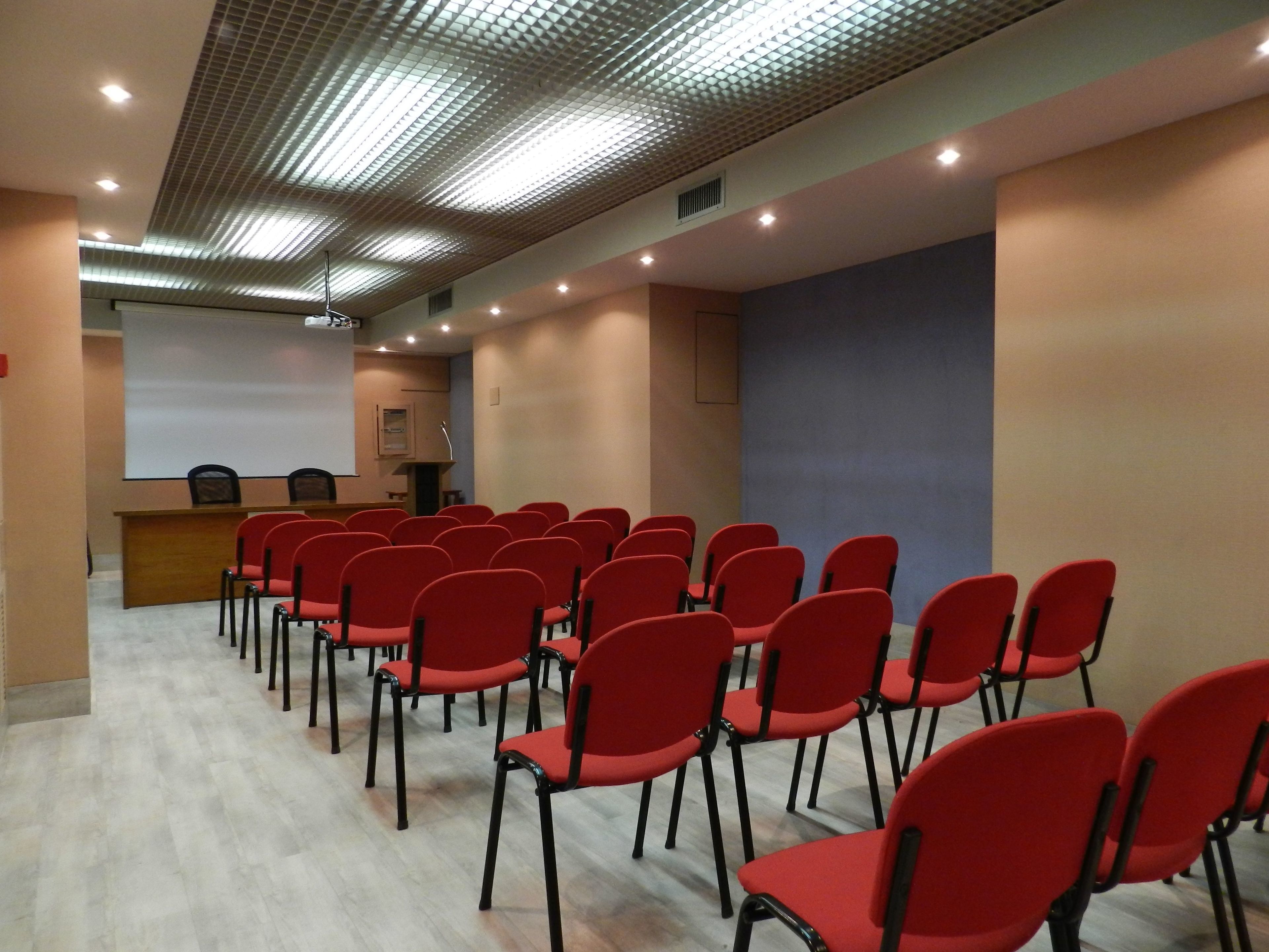 A conference room with rows of red chairs, a podium, and a projector screen.