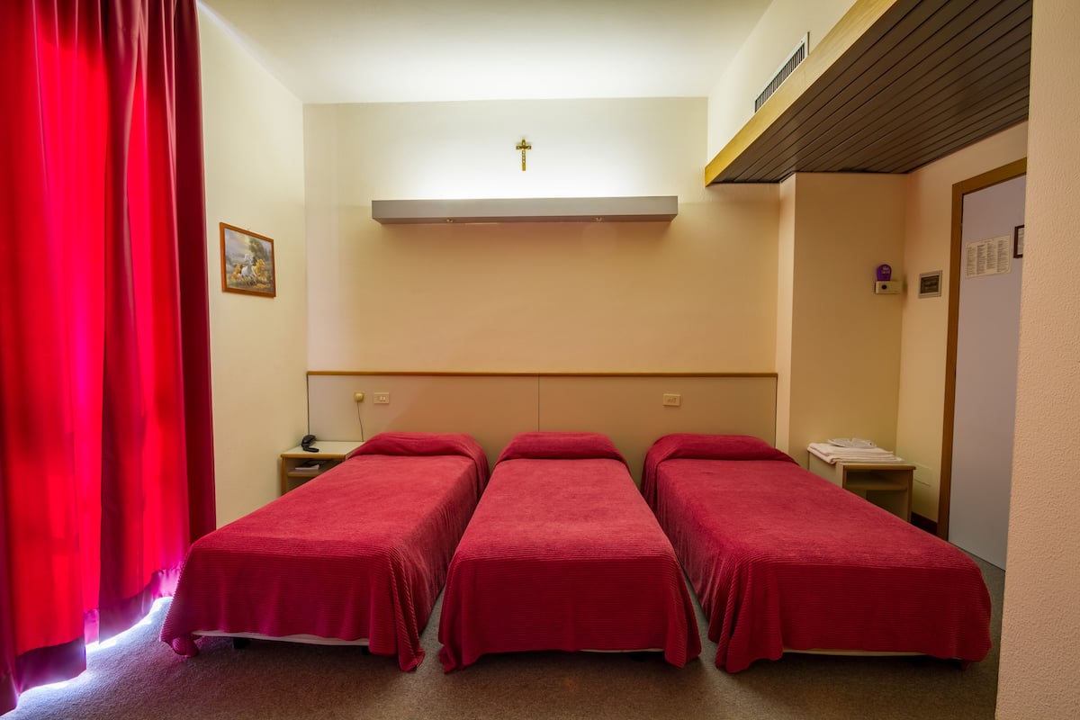 Una habitación con tres camas individuales, una mesita de noche pequeña con una lámpara, un estante en la pared con una cruz y una puerta con un tablón de anuncios.