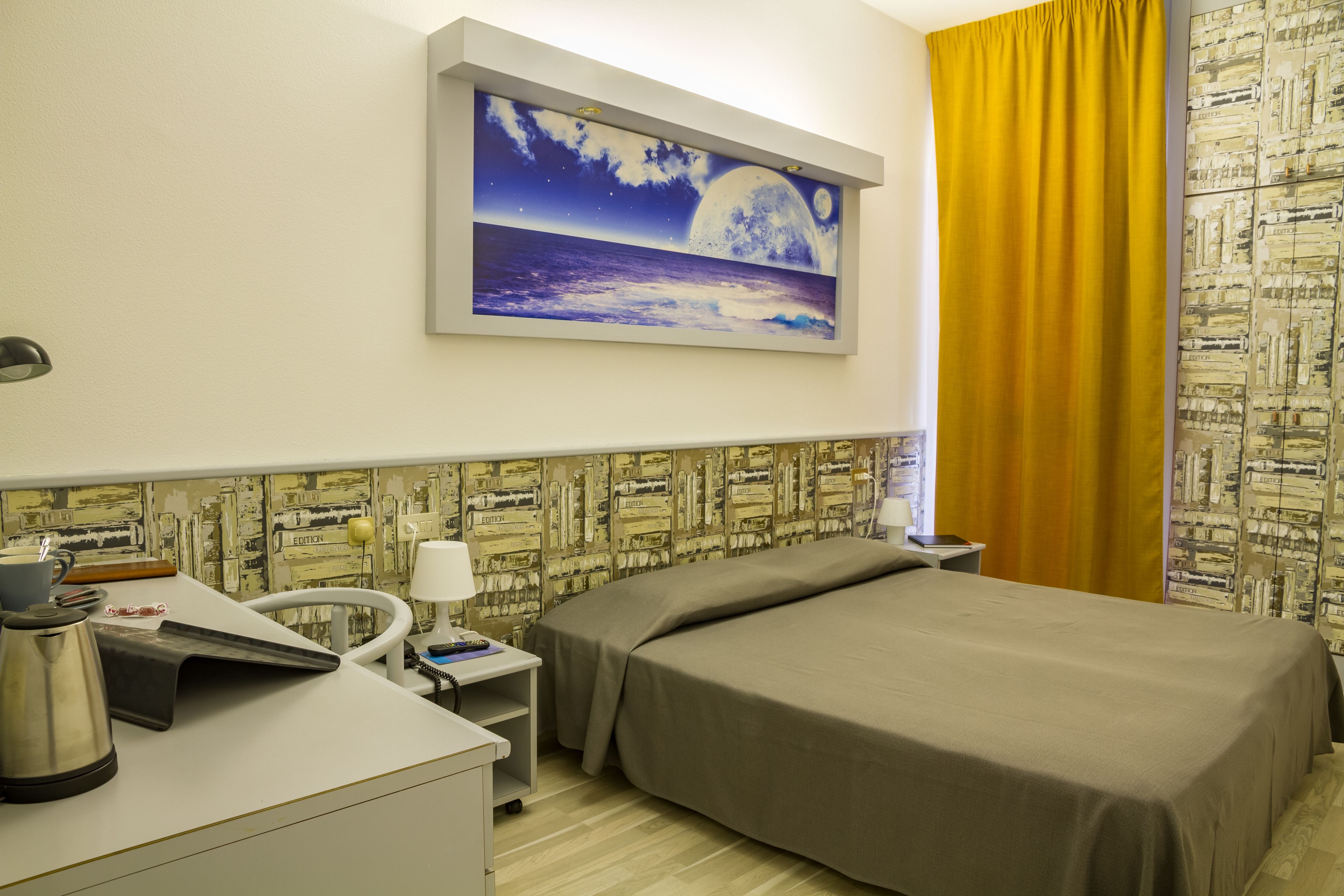 A hotel room with a bed, a desk with a kettle, a chair, a nightstand, and a painting of a night scene on the wall.