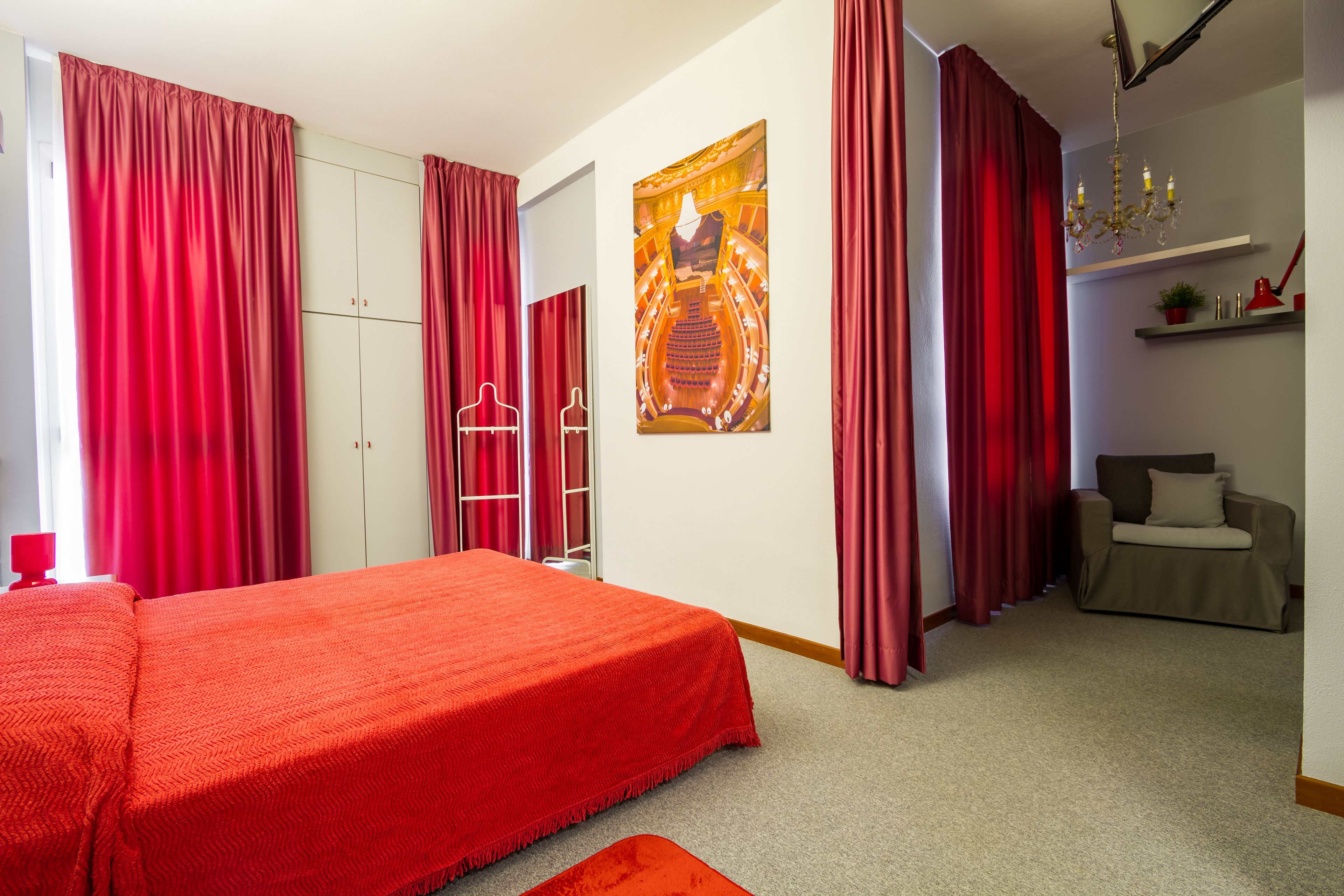Een slaapkamer met een groot bed, rode gordijnen, een kledingkast, een spiegel, een schilderij, een kroonluchter en een zithoek met een bank en een klein tafeltje.