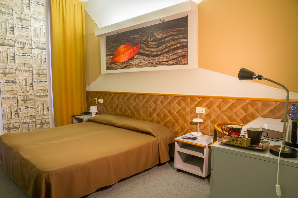 Habitación de hotel con cama, mesita de noche, escritorio, silla, lámpara, un cuadro y un papel tapiz con diseño.