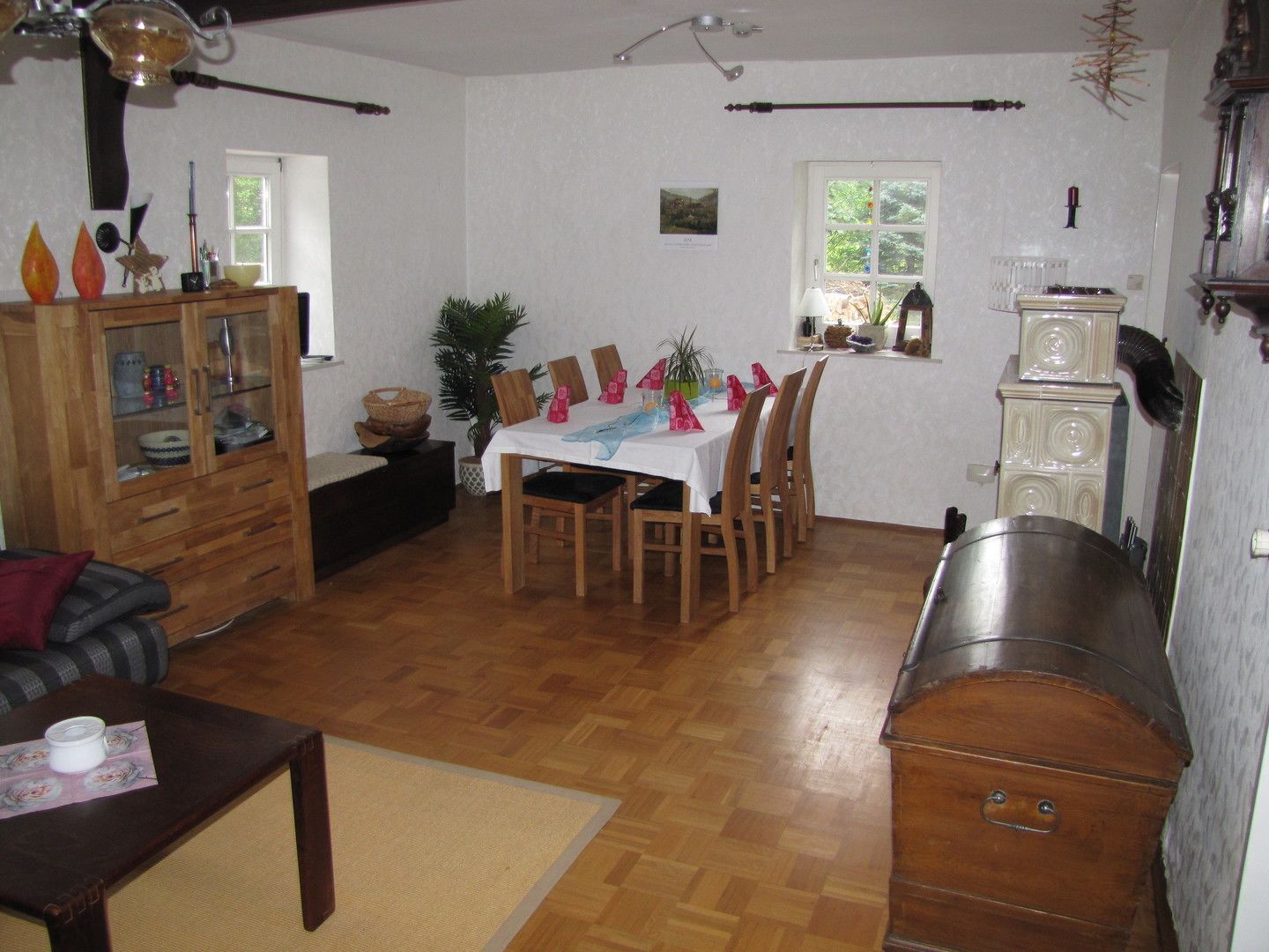 A dining area with a wooden table set for four, surrounded by chairs. There is a cabinet with glass doors to the left, a sofa with cushions, and a small table with a decorative item. A window with a view of greenery is visible in the background.