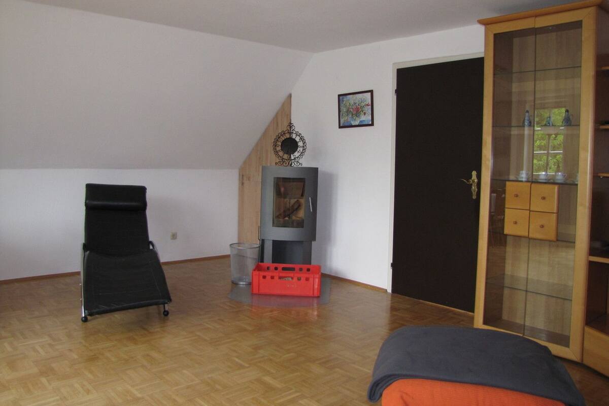 Una habitación amplia con piso de madera, una silla negra, un caja roja, una chimenea y un aparador de vidrio con objetos exhibidos.