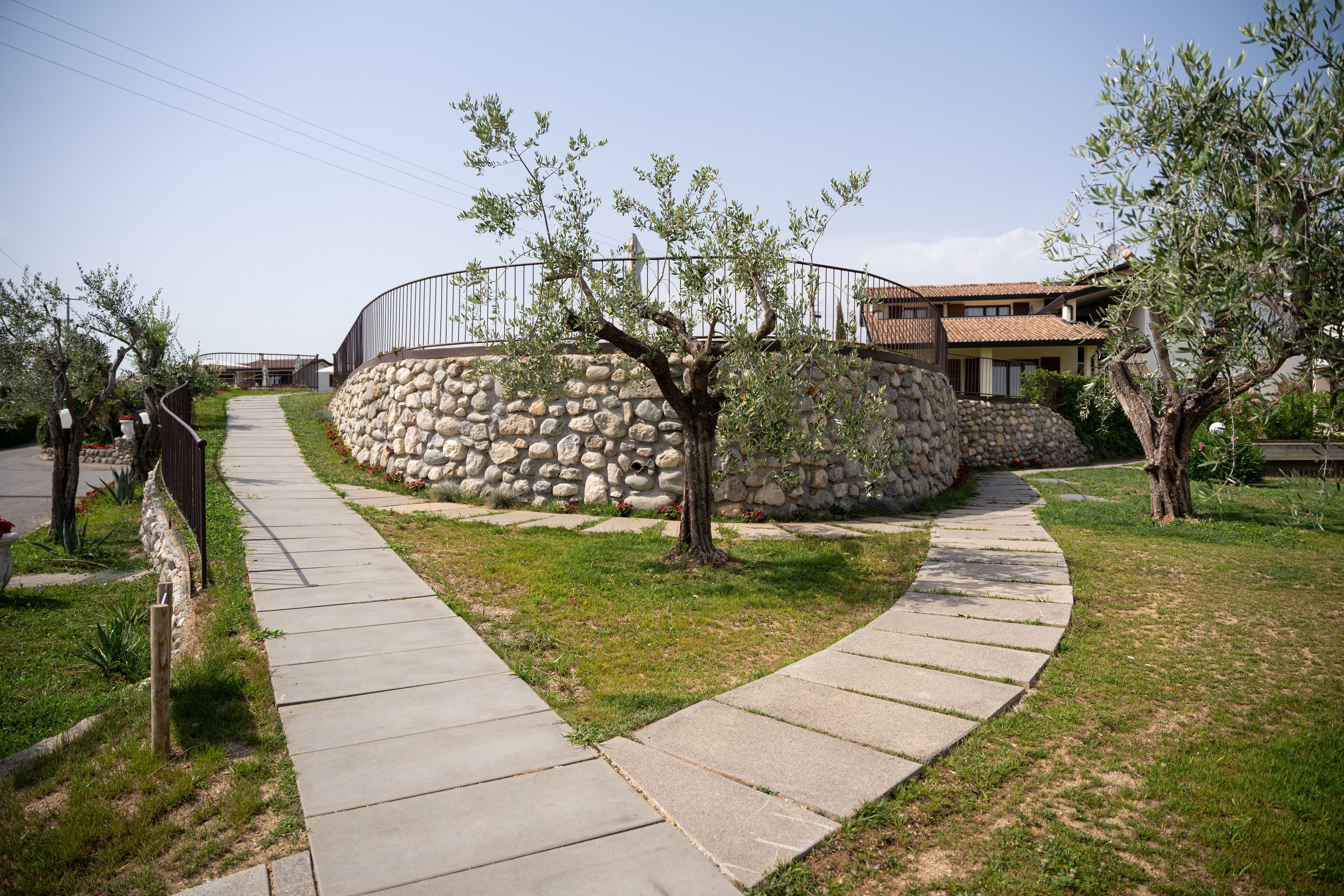 A paved pathway leading to a stone wall with a curved metal railing, surrounded by trees and grass.