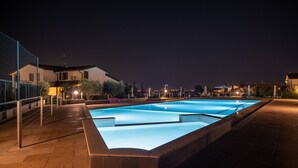 Une piscine rectangulaire, bordée d’une terrasse carrelée, entourée d’une clôture et avec quelques bâtiments visibles au loin.