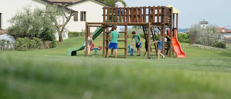 A playground with a wooden climbing structure and slides, surrounded by a grassy area and a house in the background.