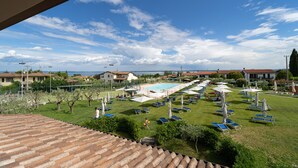 Une vue d’une zone de piscine avec plusieurs chaises longues et parasols, entourée de verdure et de bâtiments.
