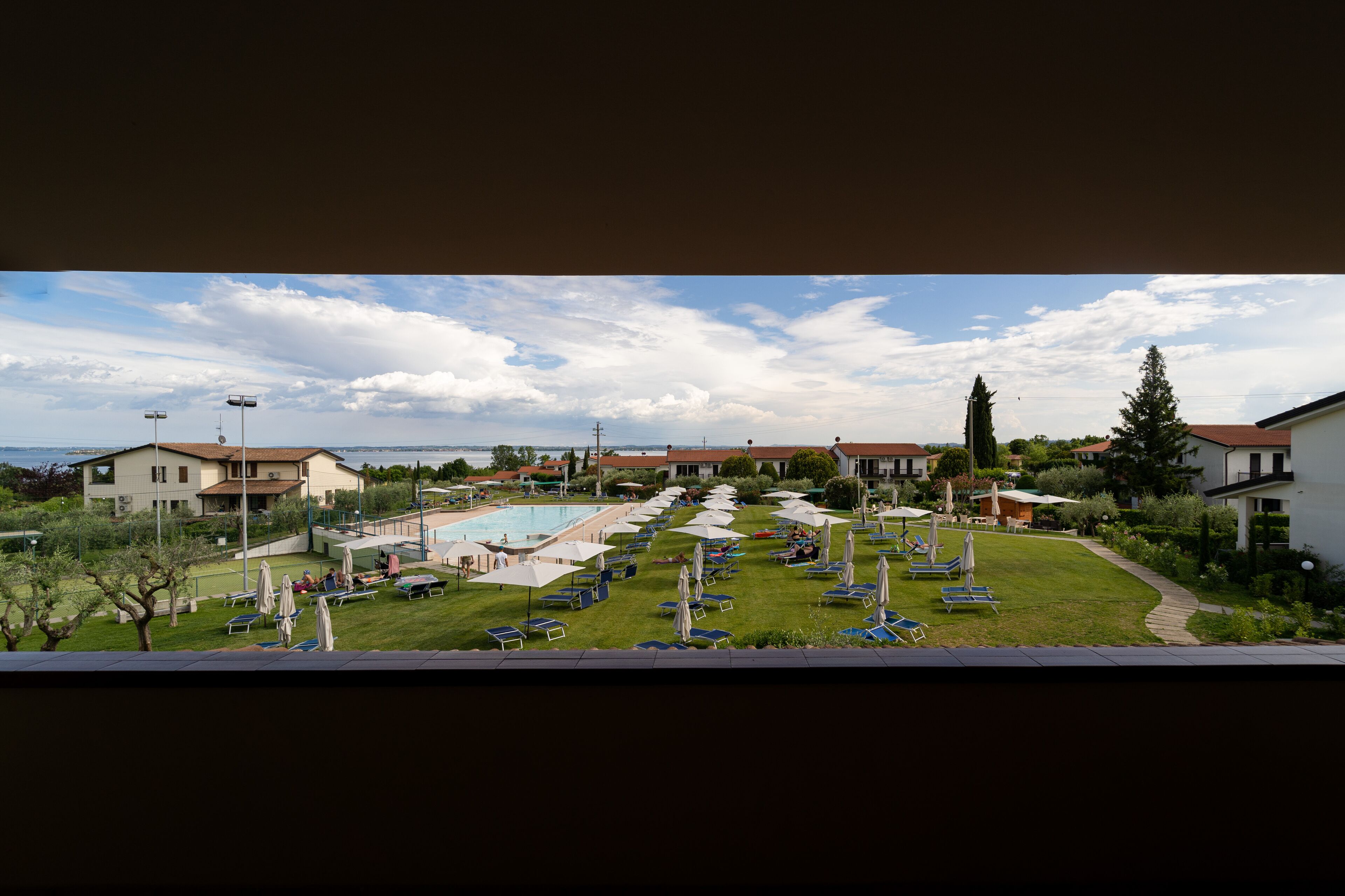 Uma área de piscina com espreguiçadeiras e guarda-sóis, cercada por edifícios e vegetação.