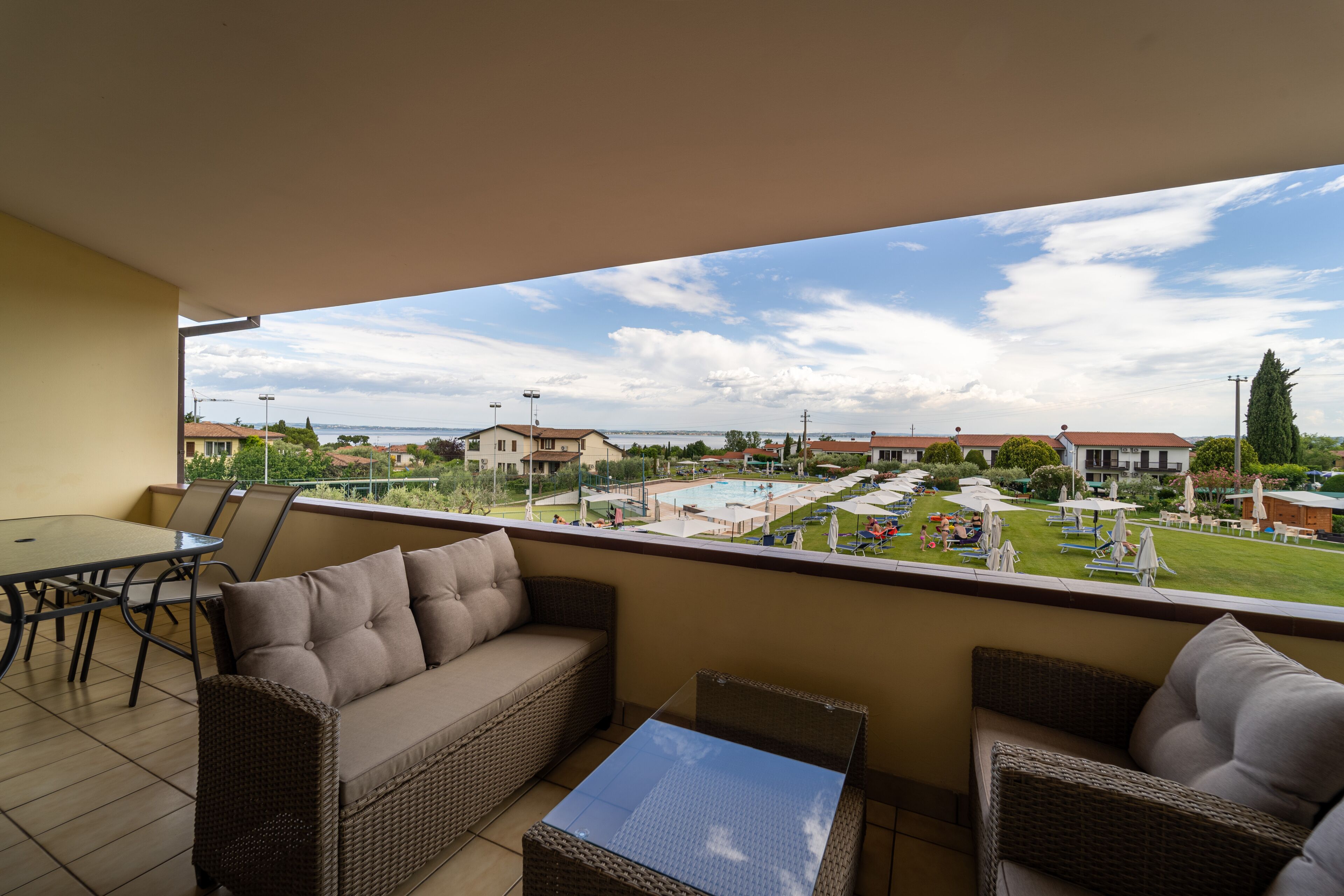 Un balcon aménagé avec des meubles en osier, une table et une vue sur un espace piscine agrémenté de parasols et de chaises longues.