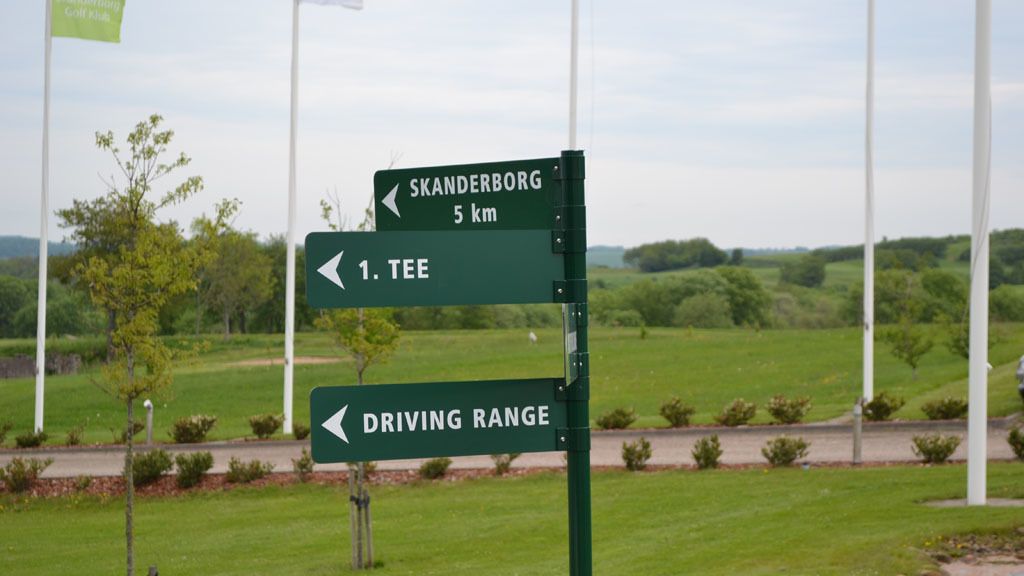 Una señal direccional con flechas que apuntan a Skanderborg y TEE, y una etiqueta que indica un campo de prácticas de golf.