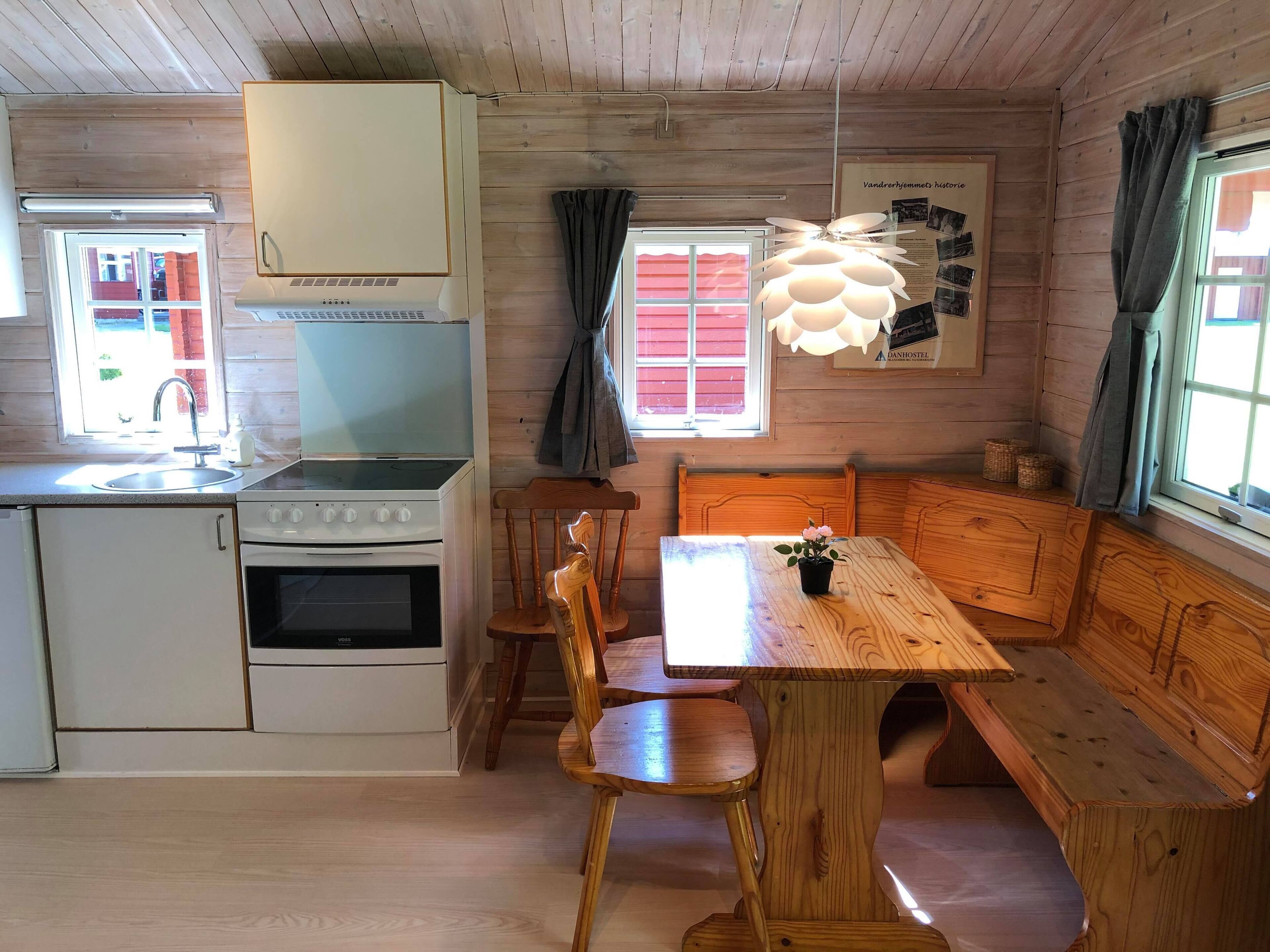 Interior de una cabaña de madera con kitchenette, mesa de comedor de madera, asientos tipo banco y una lámpara colgante.