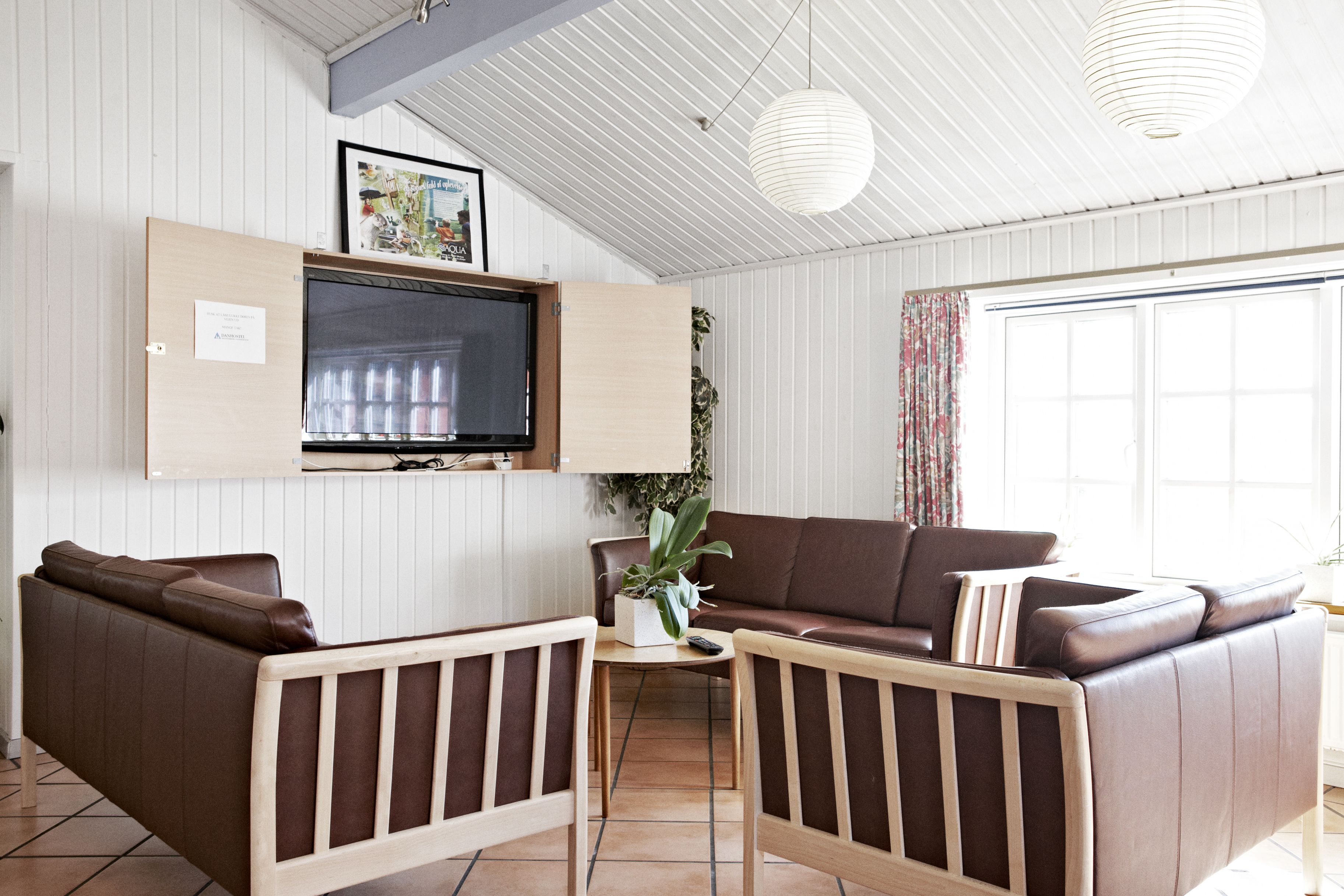 A living room with a brown leather sofa, a wooden coffee table, a TV mounted on the wall, and a large window with curtains.