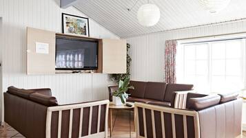 A living room with a brown leather sofa, a wooden coffee table, a TV mounted on the wall, and a large window with curtains.