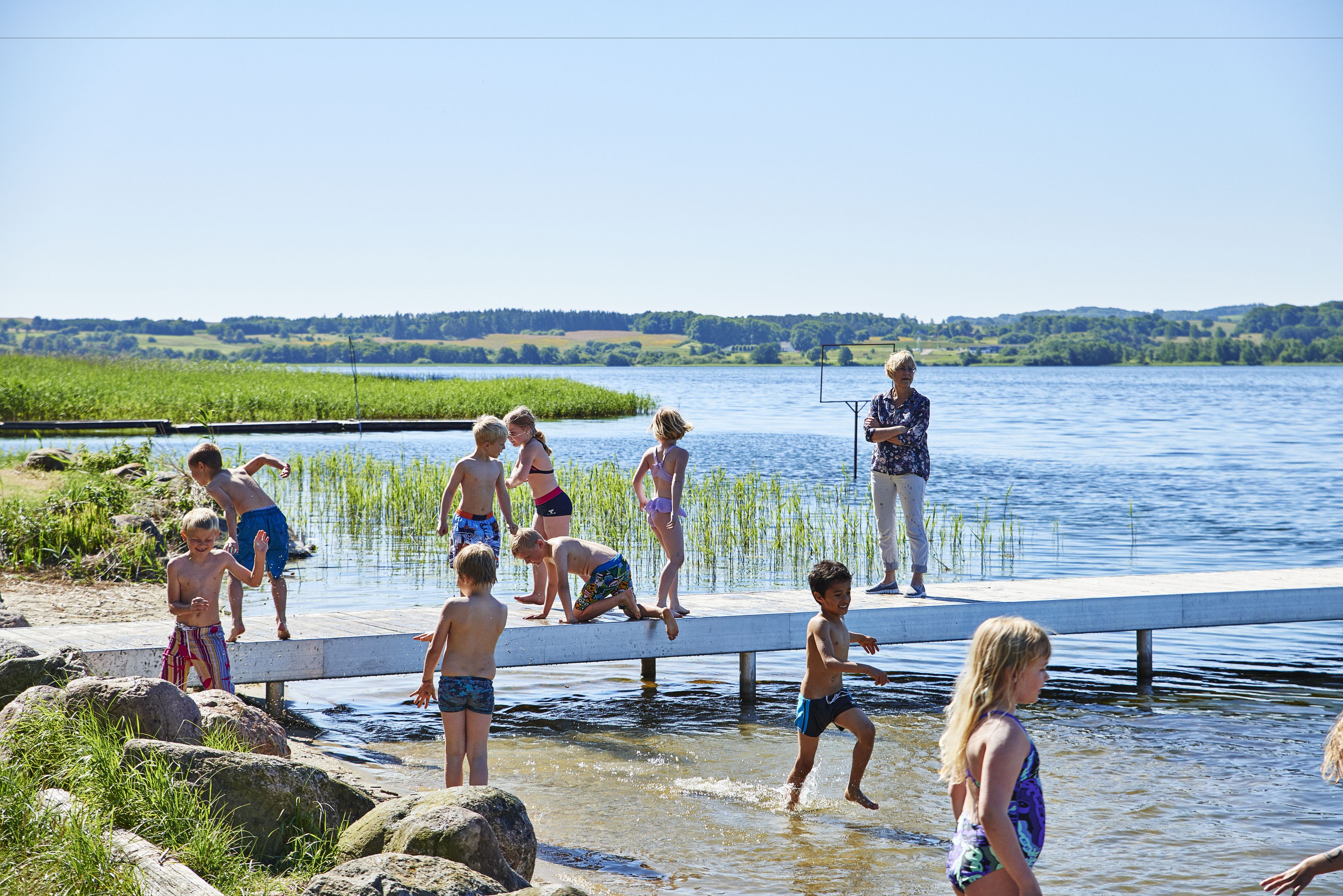 Un groupe de personnes se trouve dans une zone de baignade au bord d’un lac, avec une jetée et une personne qui s’y tient debout.