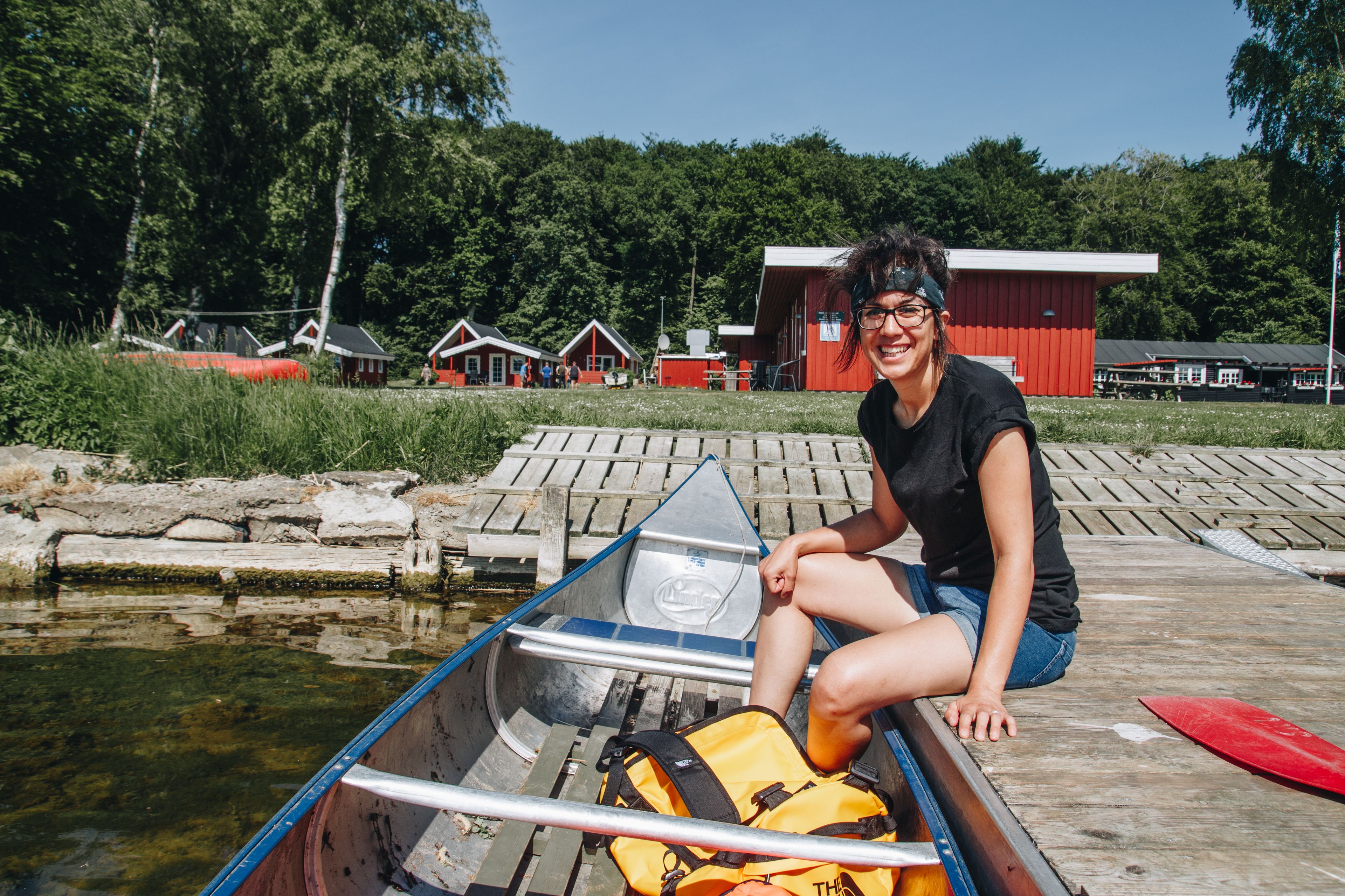 A person sitting in a canoe on a wooden dock with a red building in the background.