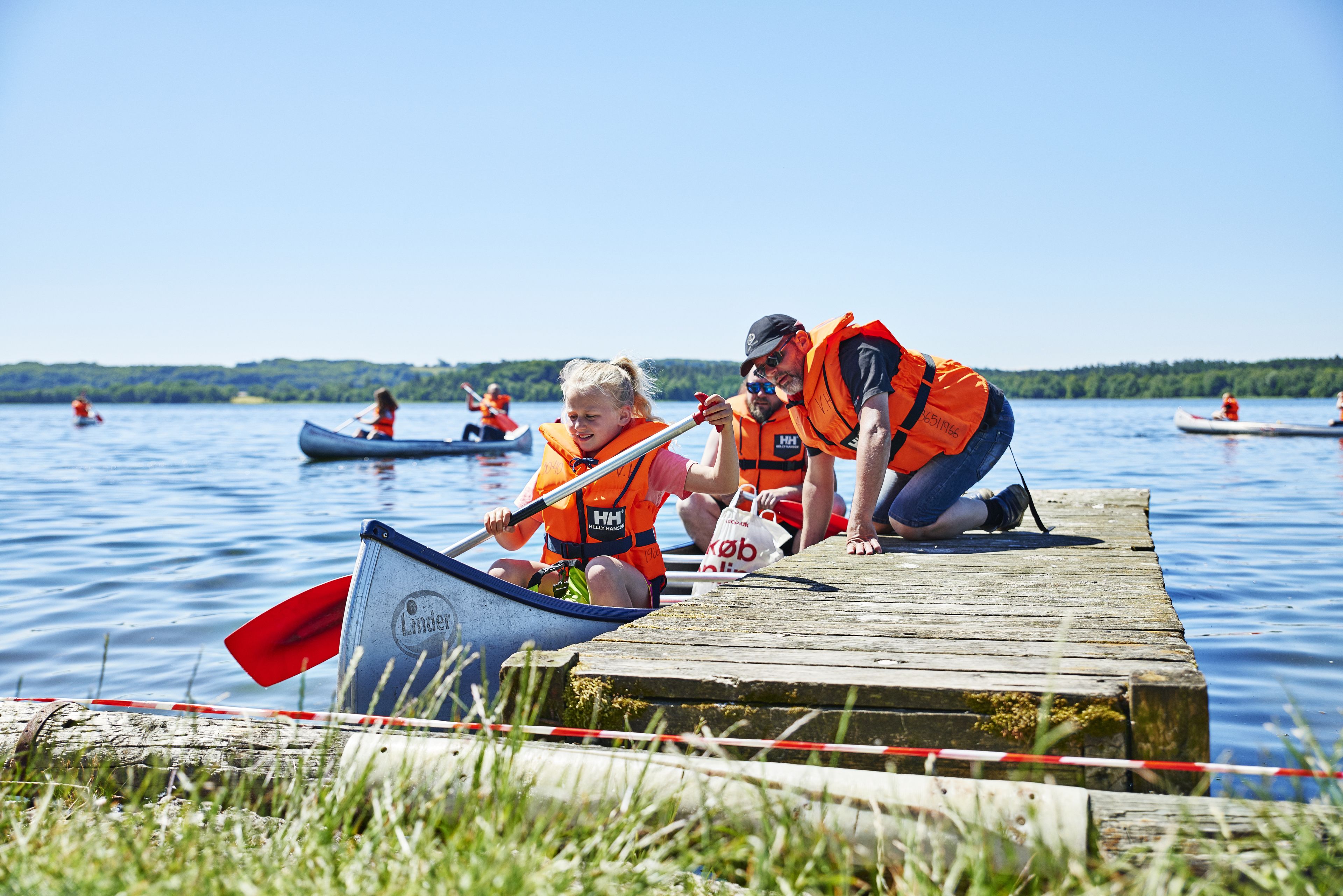 Un muelle de madera que se adentra en un lago, con gente en canoas y kayaks.