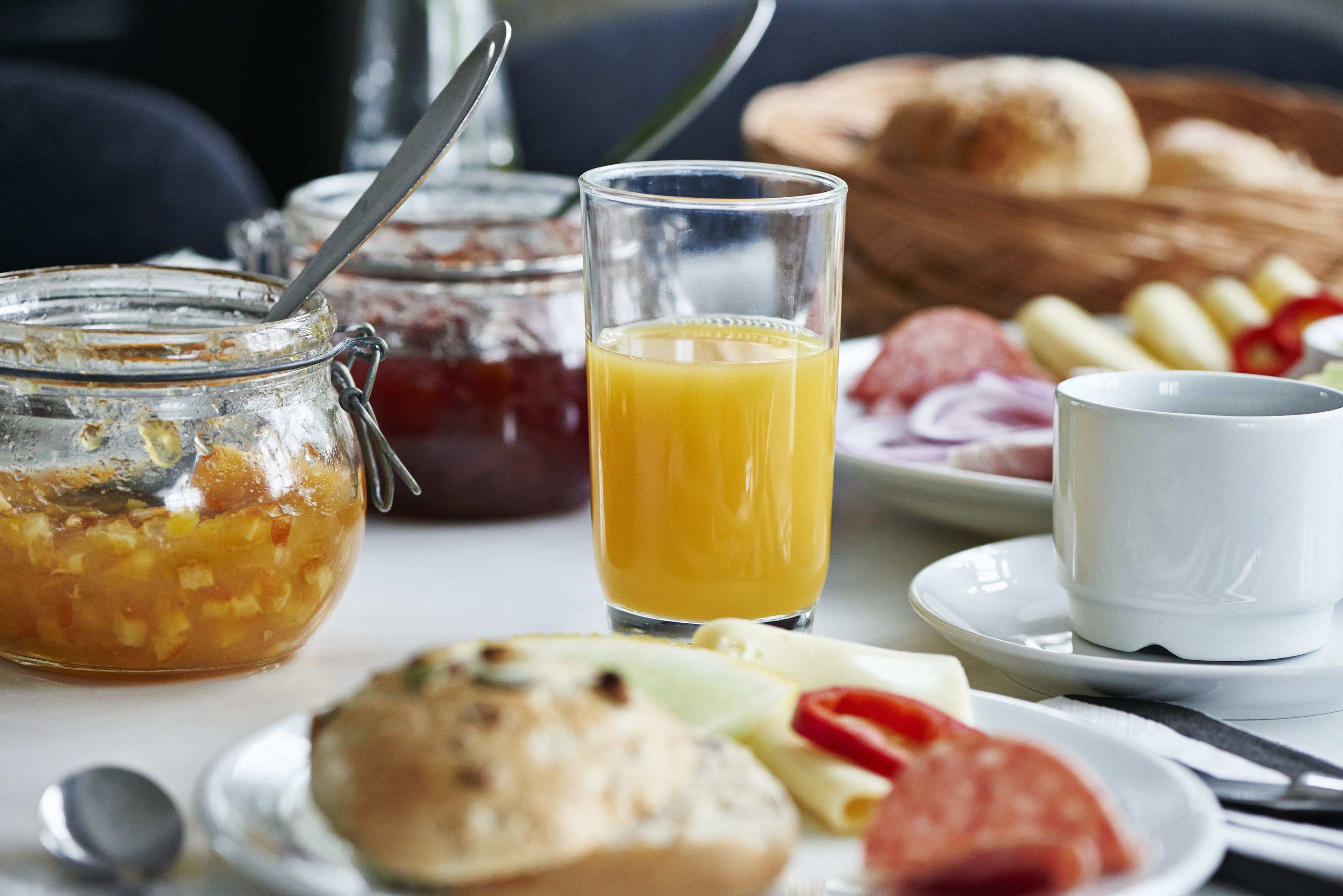 Un desayuno con un vaso de jugo de naranja, un frasco de mermelada, un plato de pan, queso y tomates, y una taza blanca sobre una mesa.
