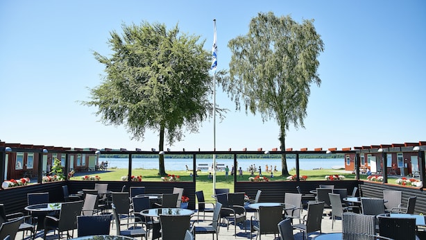 Een terras met tafels en stoelen, met uitzicht op een waterpartij en een gazon met mensen.