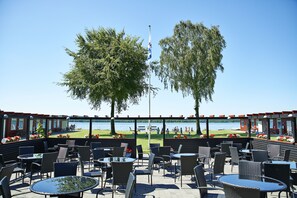 An outdoor seating area with tables and chairs, overlooking a body of water and a grassy area with people. - Danhostel Skanderborg (Skanderborg)