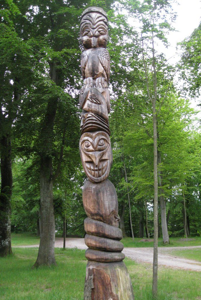 A totem pole with intricate carvings, set in a forested area with a dirt path in the background.