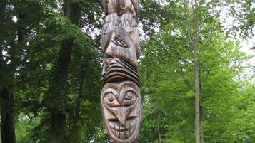 A totem pole with intricate carvings, set in a forested area with a dirt path in the background.
