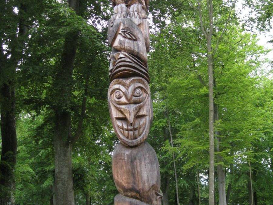 A totem pole with intricate carvings, set in a forested area with a dirt path in the background.