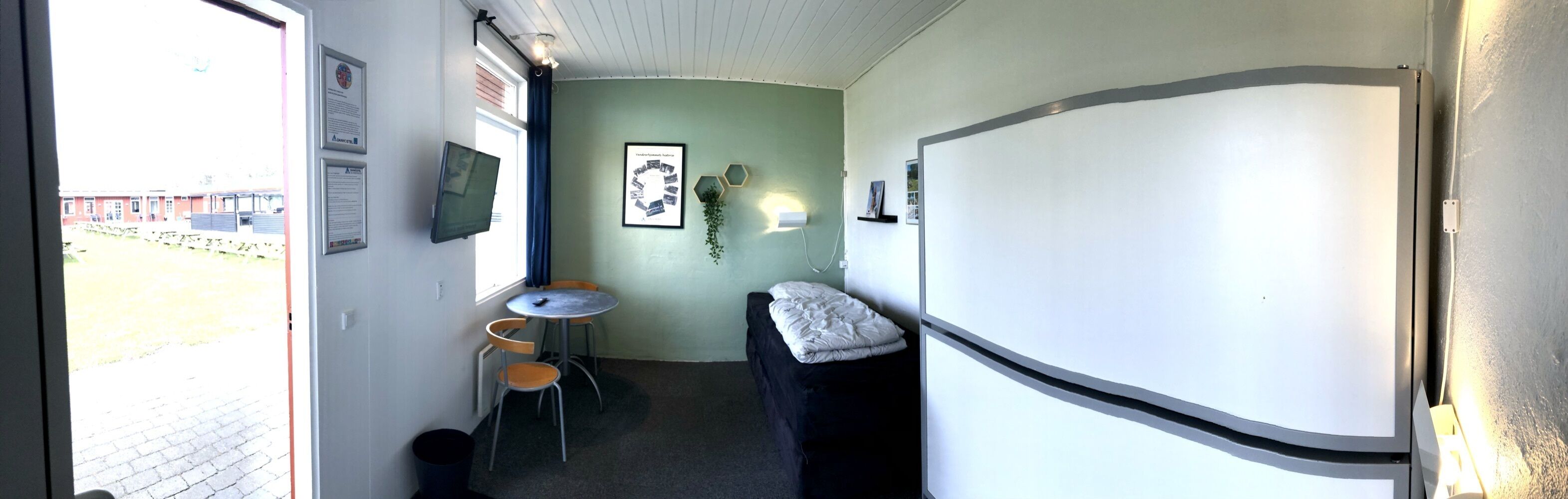 A small, narrow room with a bed, a table, and a chair. There are framed pictures on the wall and a window with a view of the outside.