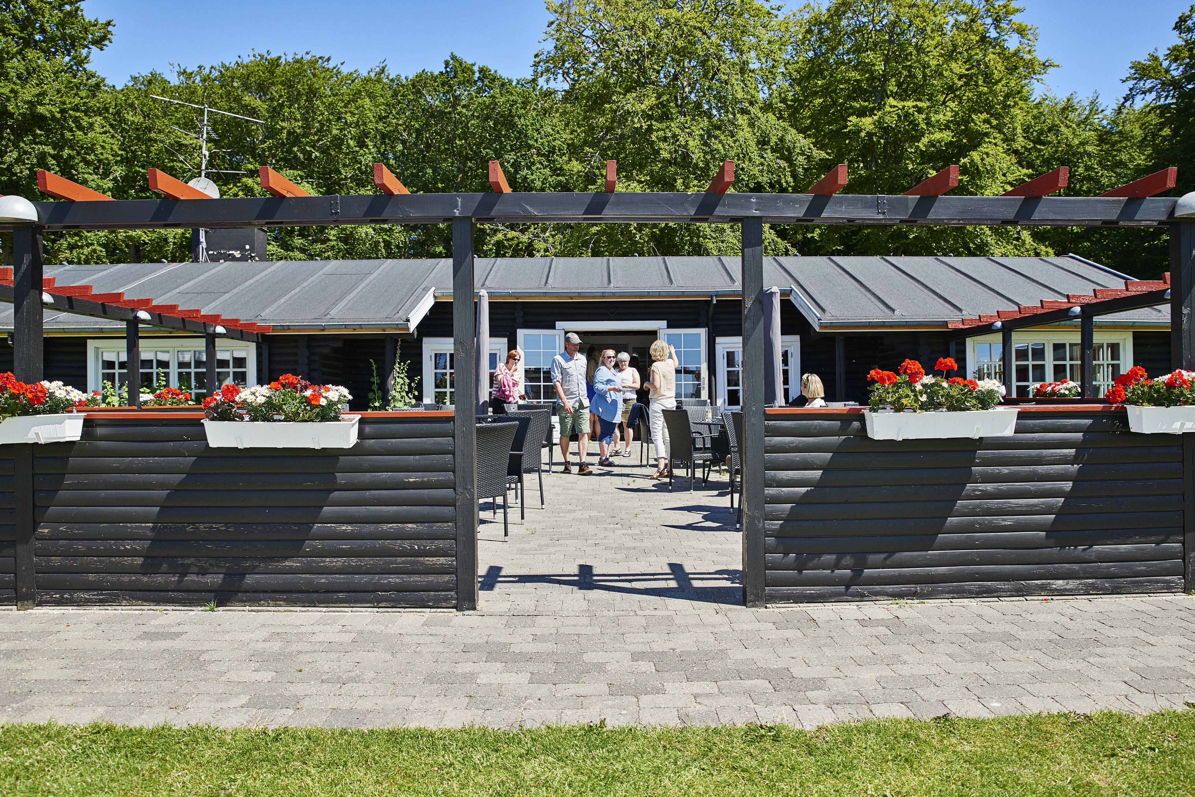 A covered outdoor area with a wooden pergola, tables, and chairs, leading to a building with a dark roof and windows.