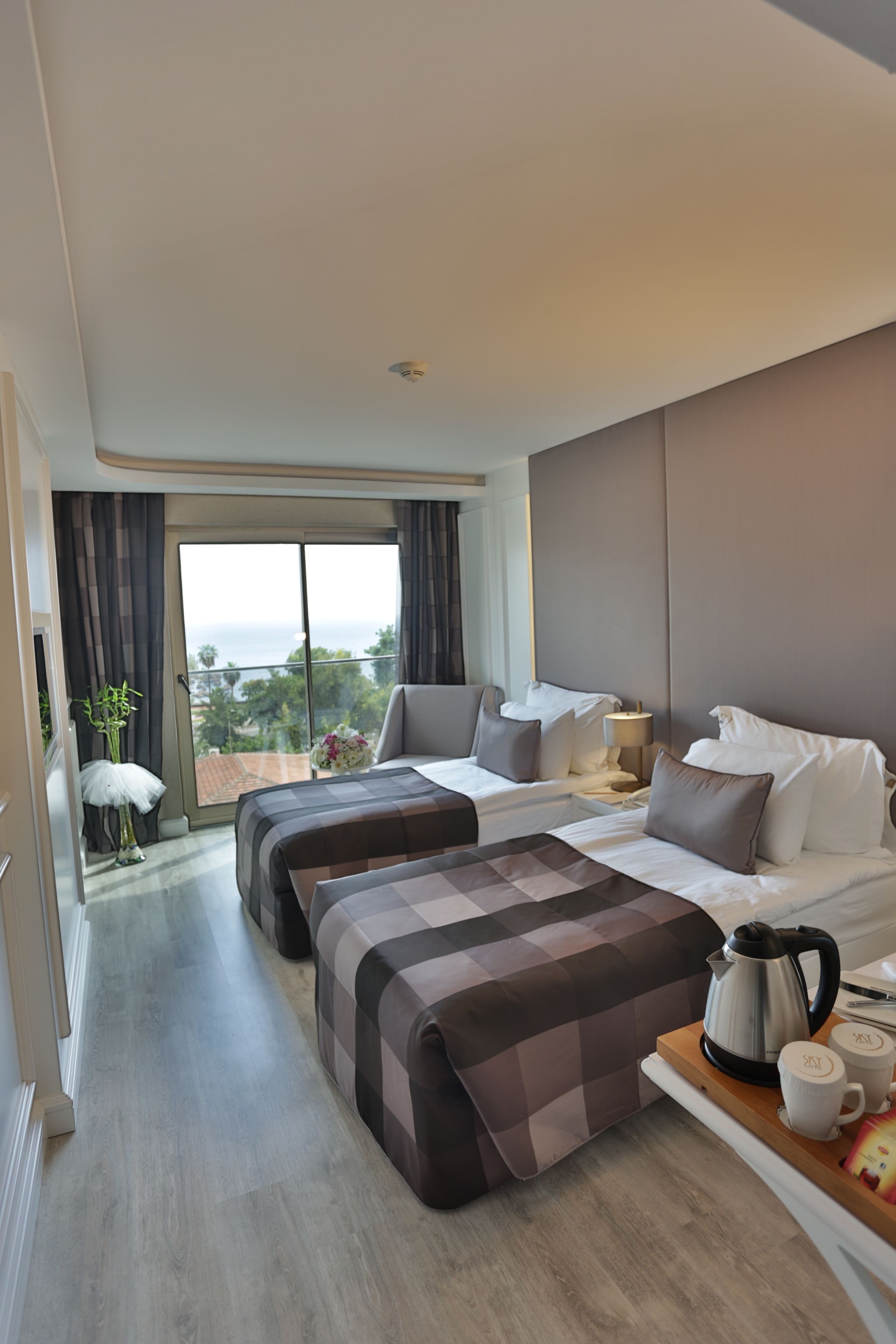 A modern hotel room with a large bed, a second smaller bed, a balcony with a view, and a coffee table with a kettle and mugs.