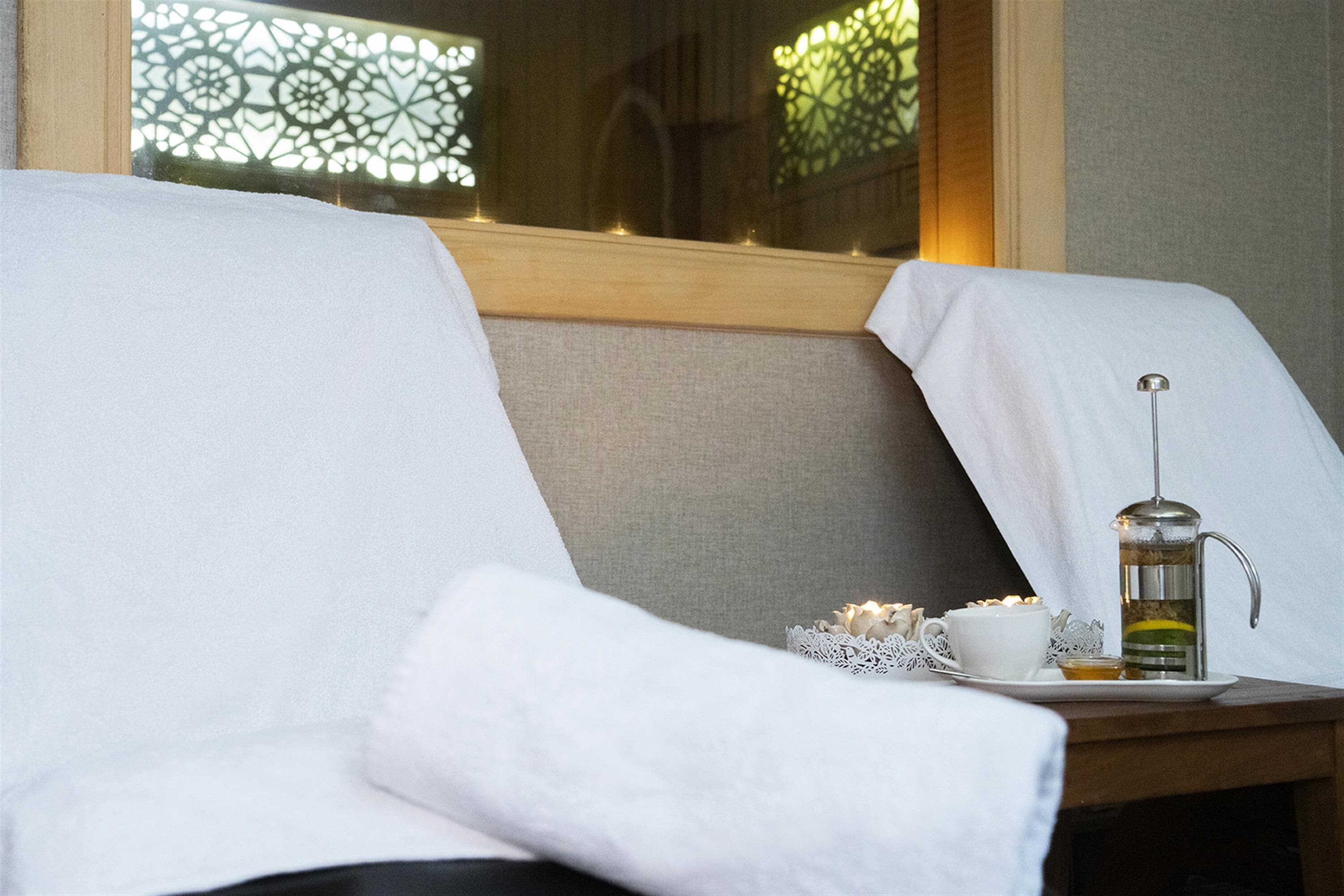 A neatly made bed with white linens, a bedside table with a teapot and cups, and a decorative window with intricate patterns.