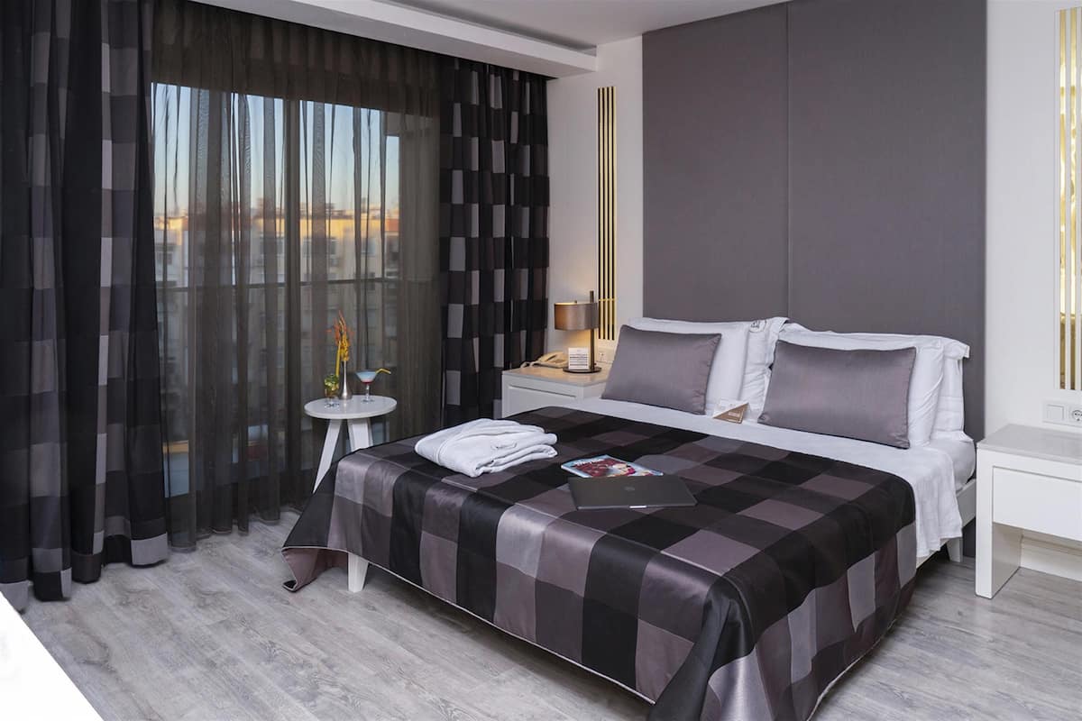 Une chambre d’hôtel moderne avec un grand lit, une table de chevet avec une lampe et une vue sur la ville à travers de fines rideaux.