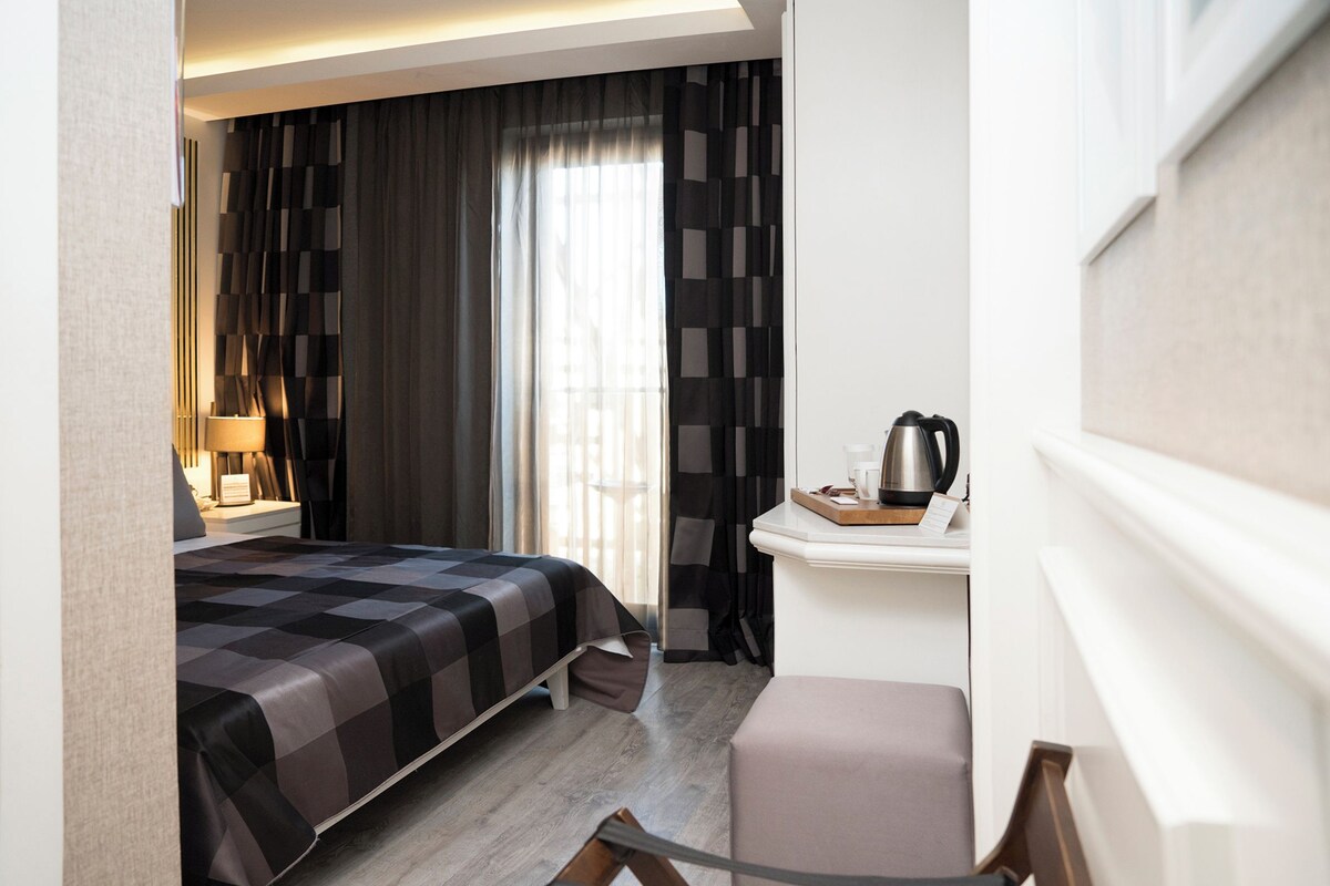 A hotel room with a bed, a desk with a kettle, a chair, and a window with curtains.