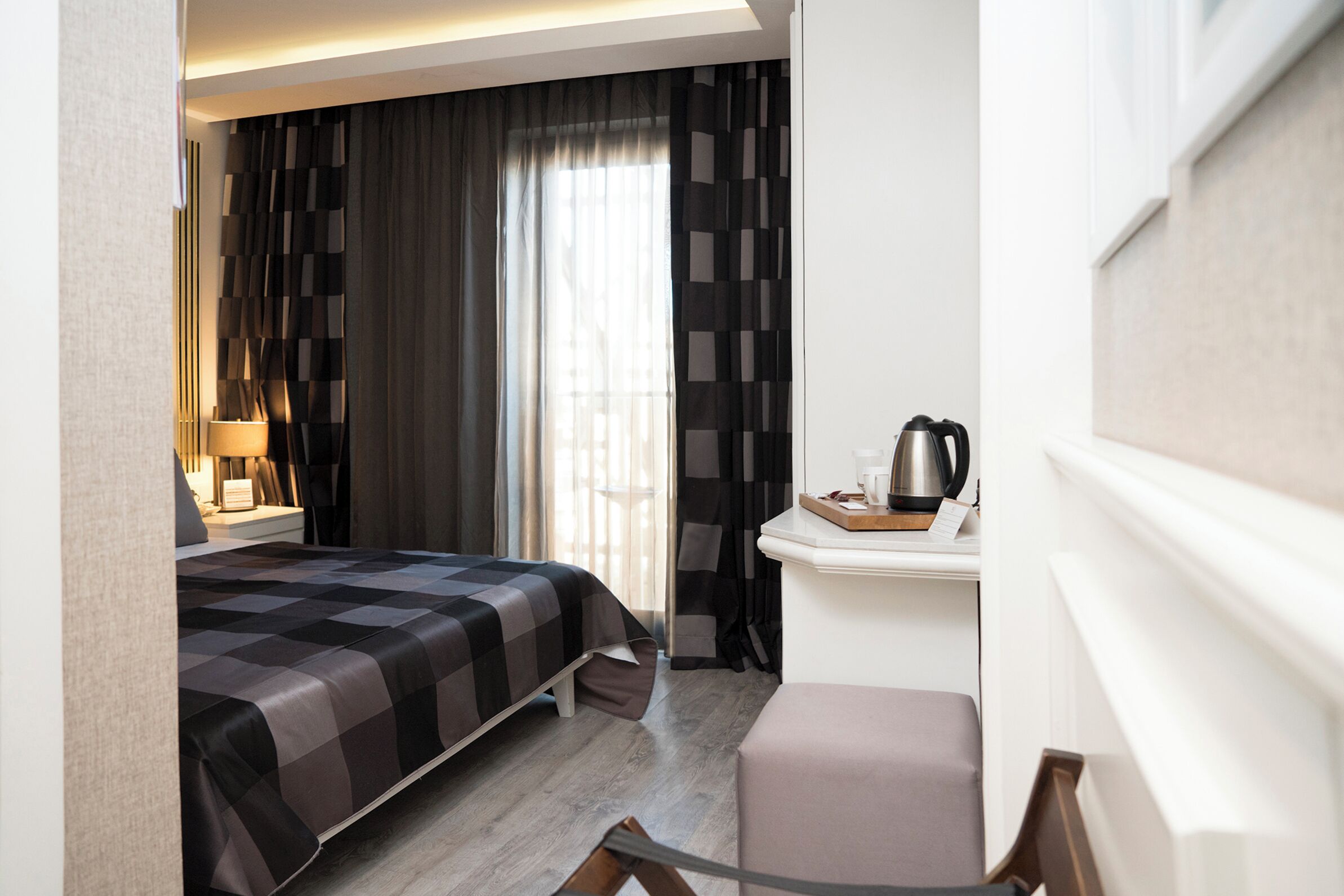 Quarto de hotel com cama, escrivaninha com chaleira, cadeira e janela com cortinas.