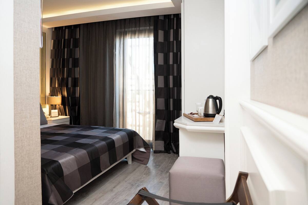 Quarto de hotel com cama, escrivaninha com chaleira, cadeira e janela com cortinas.