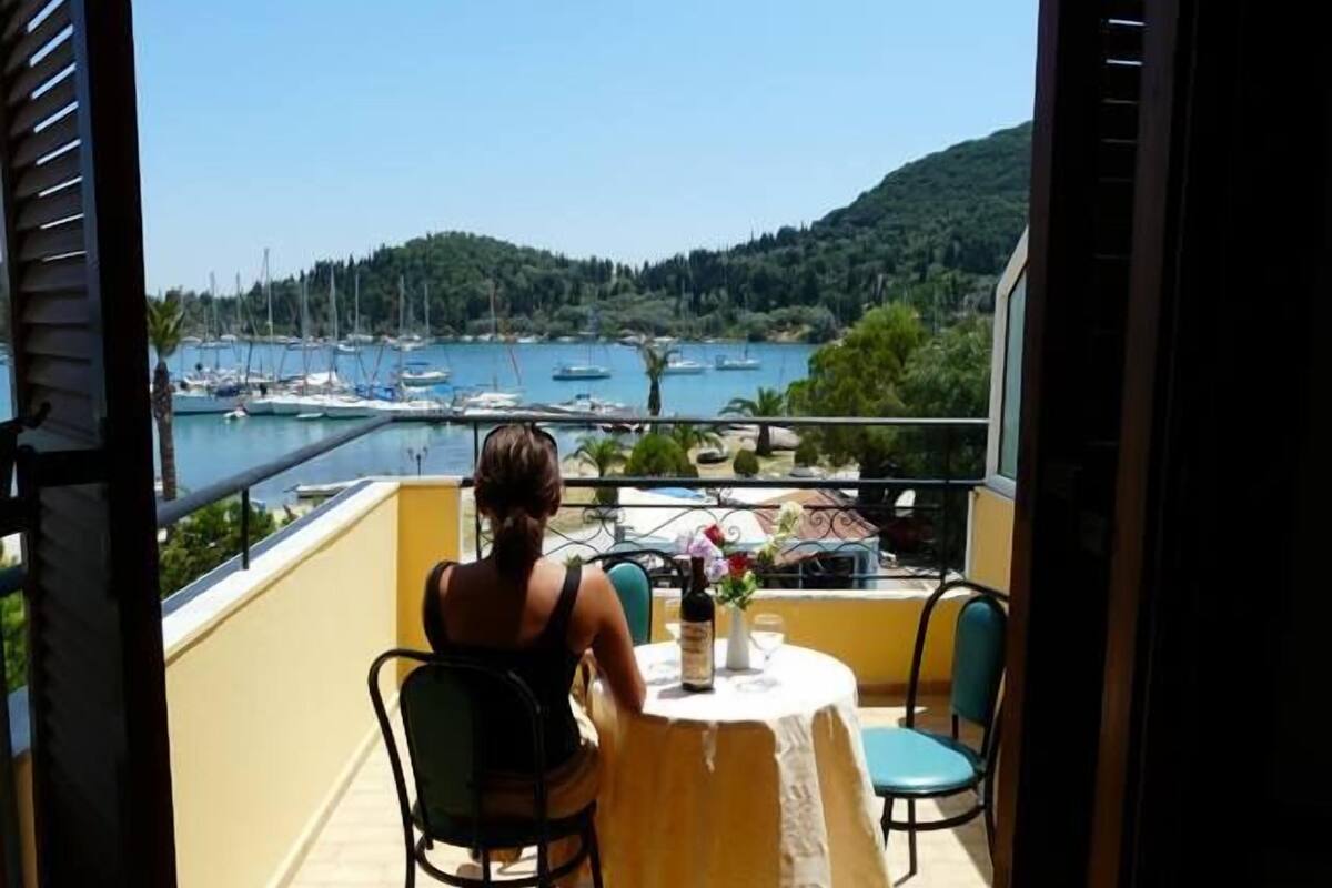 Un balcón con mesa y sillas que da a un puerto deportivo con barcos y un paisaje montañoso.