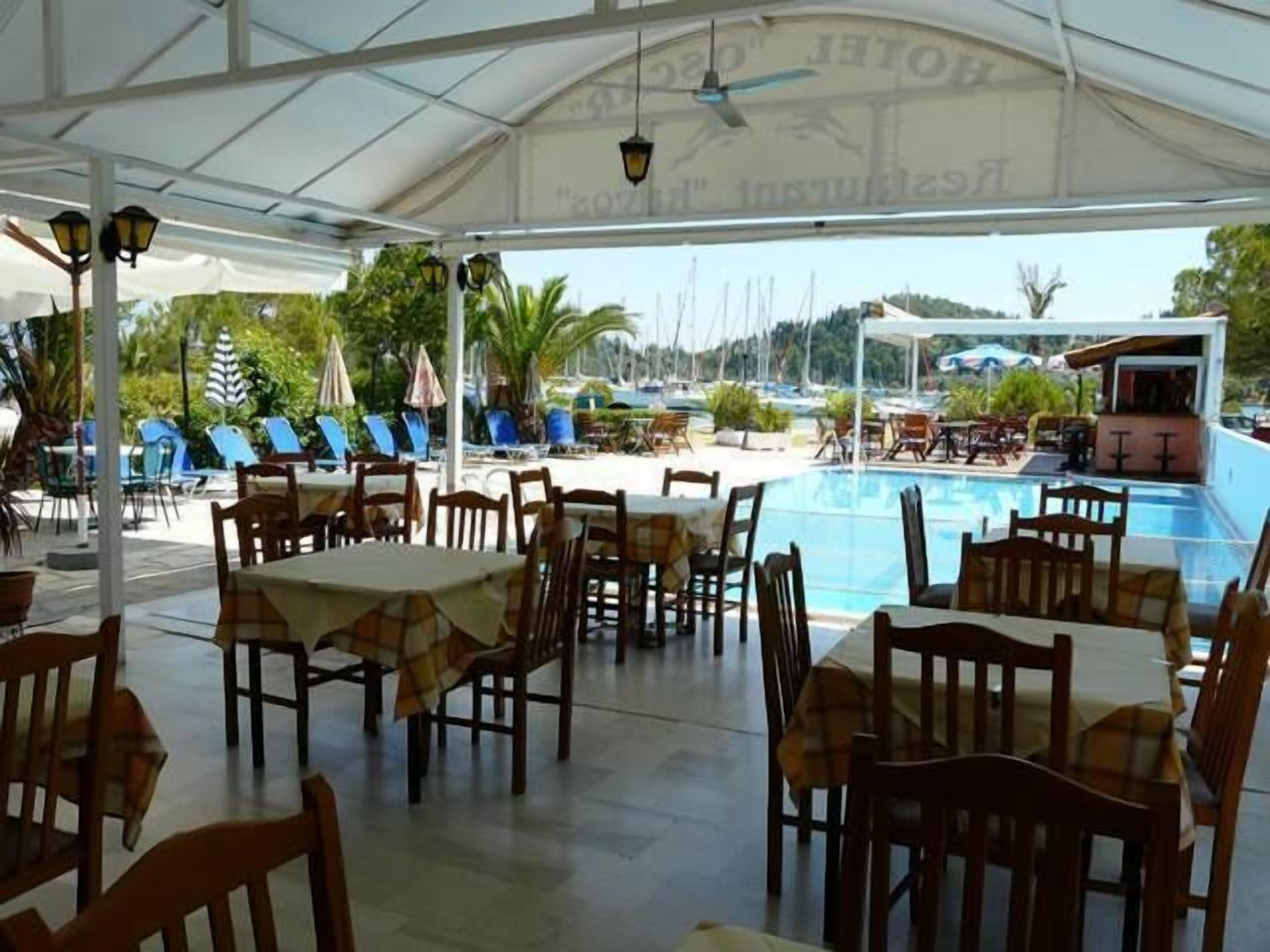 Uma área de refeições coberta ao ar livre, com mesas e cadeiras, com vista para uma piscina e um porto com barcos.