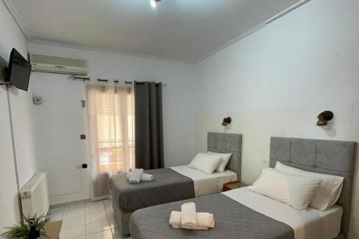 Habitación de hotel con dos camas, una mesita con una botella y vasos, un televisor montado en la pared y una ventana con cortinas.