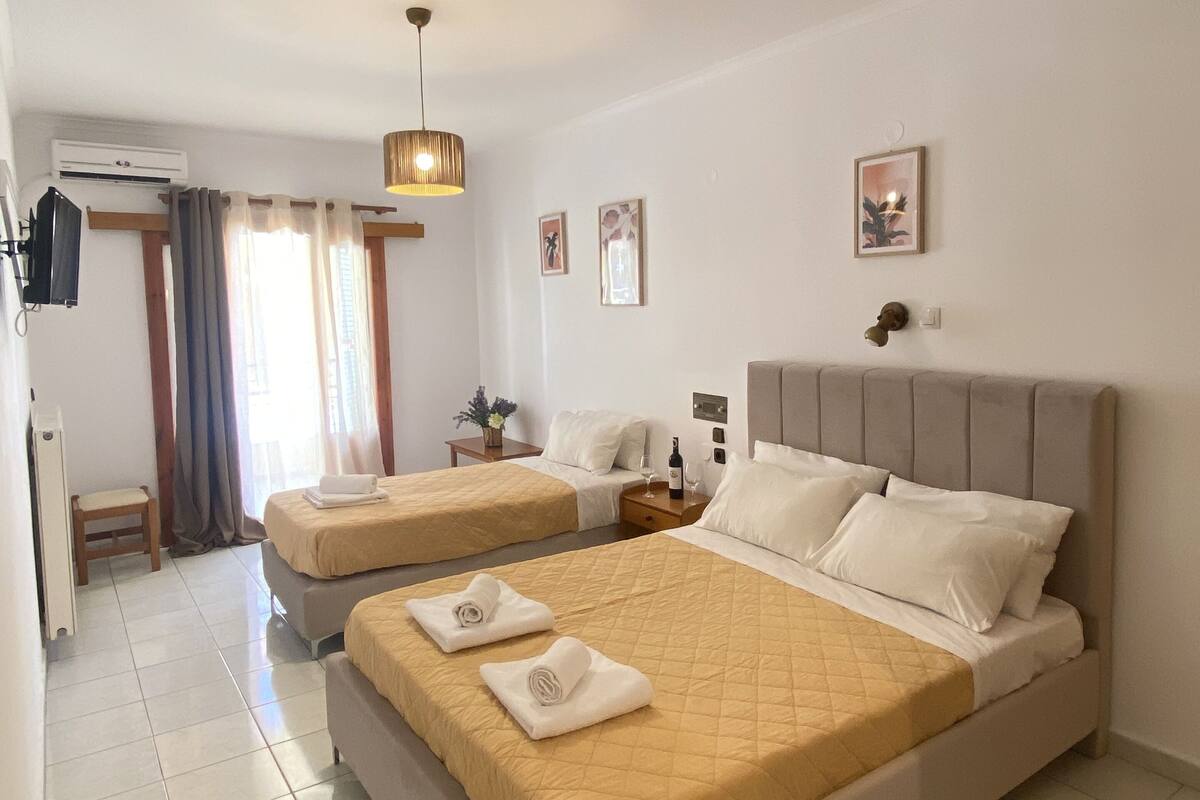 Una habitación de hotel con dos camas, una mesita de noche con una botella y vasos, una lámpara colgante y un pequeño taburete.