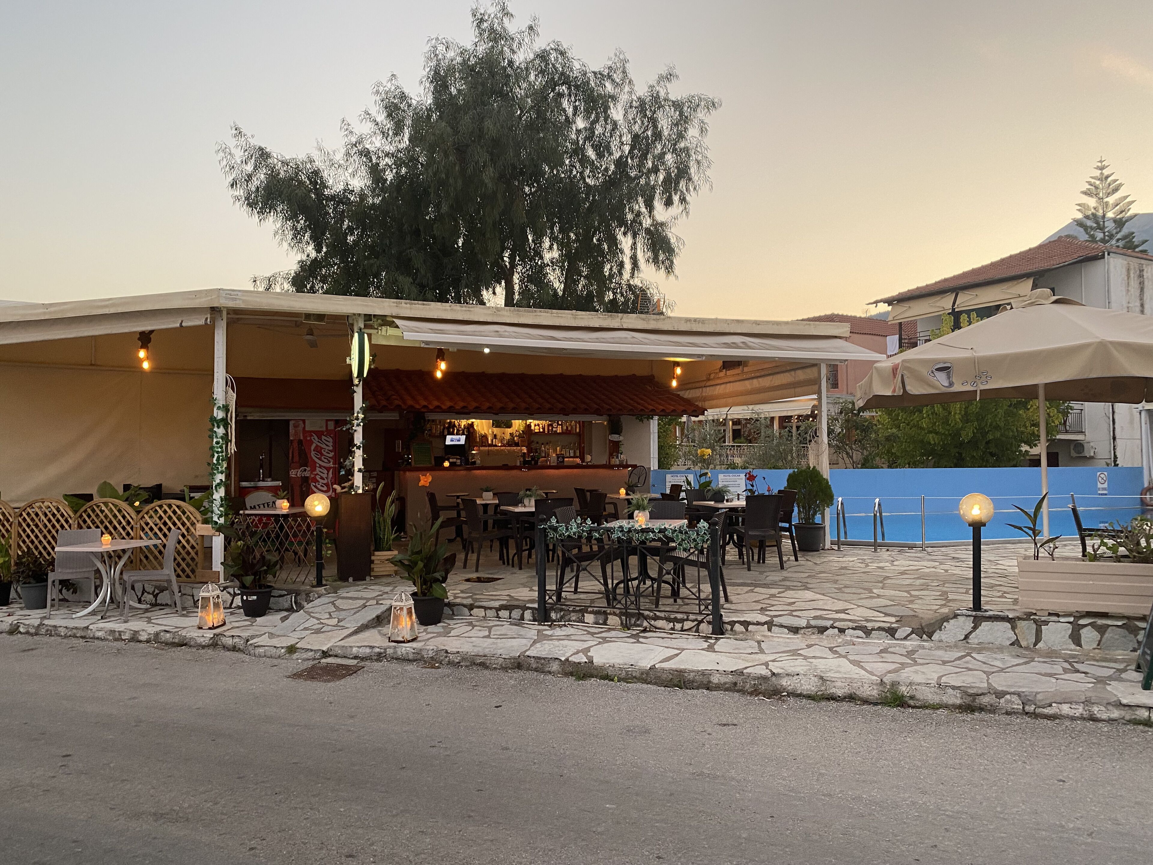 An outdoor café with tables and chairs, a bar area, and a large tree in the background.