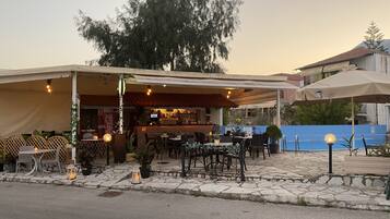 An outdoor café with tables and chairs, a bar area, and a large tree in the background.