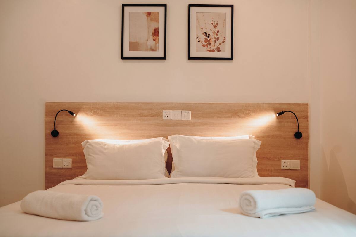 A neatly made bed with white linens, two wall-mounted light fixtures, and two framed artworks above it.