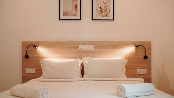 A neatly made bed with white linens, two wall-mounted light fixtures, and two framed artworks above it.