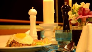 Una mesa con una vela, saleros y pimenteros, un arreglo floral, un plato con un huevo frito y fideos, y vasos de agua y té.