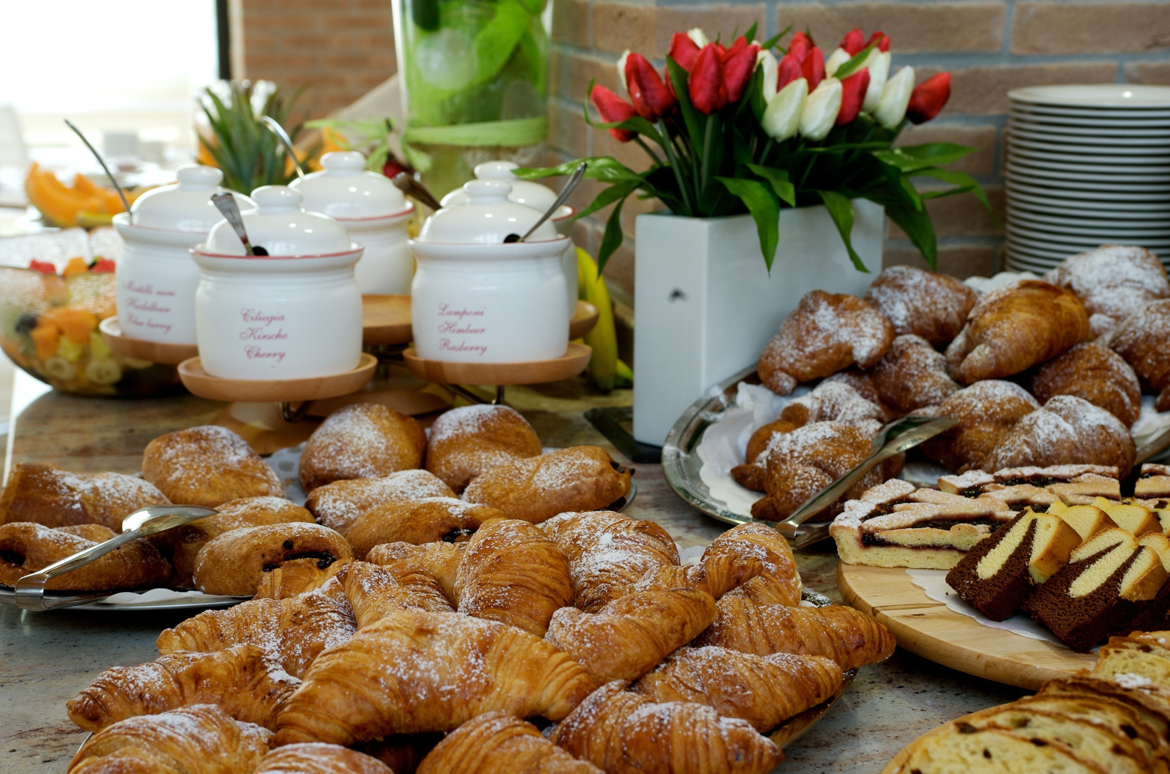 A buffet spread with various pastries, including croissants and cakes, and a vase of red and white tulips.