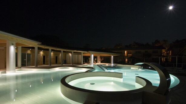 A nighttime swimming pool area with a circular design, surrounded by covered seating areas and a building with multiple windows. - Green Village Eco Resort (Lignano Sabbiadoro)
