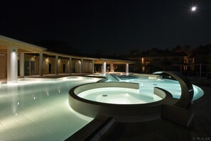 Area piscina notturna con un design circolare, circondata da zone relax coperte e un edificio con numerose finestre.