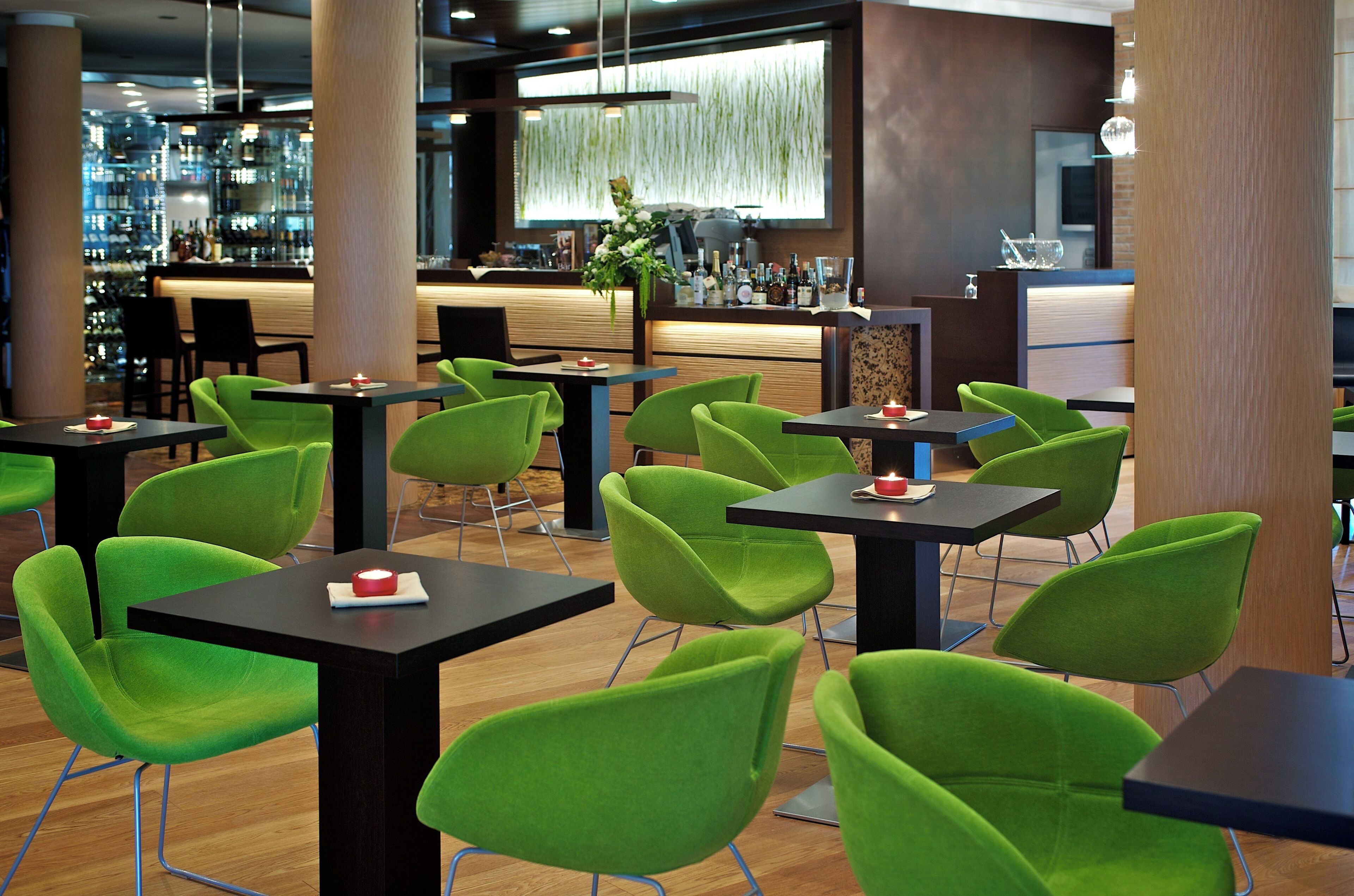 A modern restaurant with green chairs and black tables, a bar area in the background, and a decorative hanging plant.